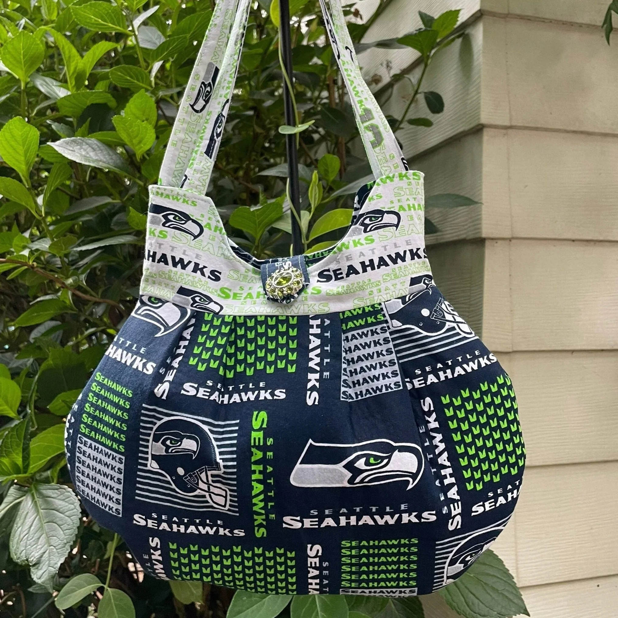 A fabric bag featuring the Seattle Seahawks football team logo and colors, hanging from a plant next to a house wall.