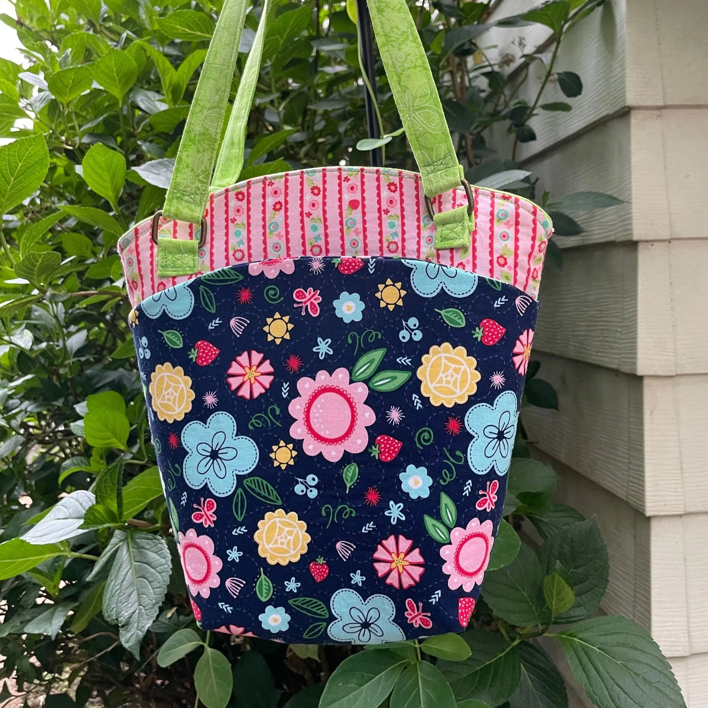Two patterned tote bags hanging on a hook among green leafy plants, with a house wall in the background. One bag is pink with red and white striped vertical lines, and another is dark blue with multicolored flowers and strawberries.