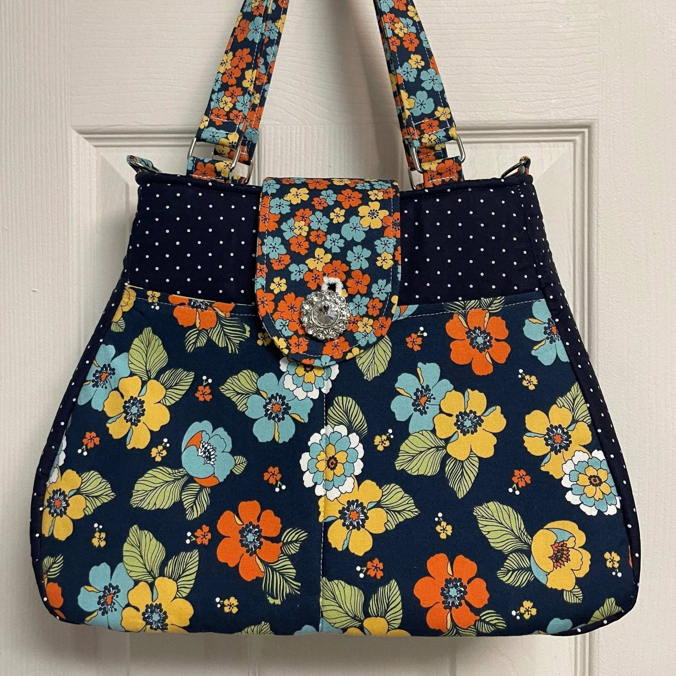 A floral-patterned fabric handbag with a flap closure featuring a decorative button and a strap, hanging on a white door.