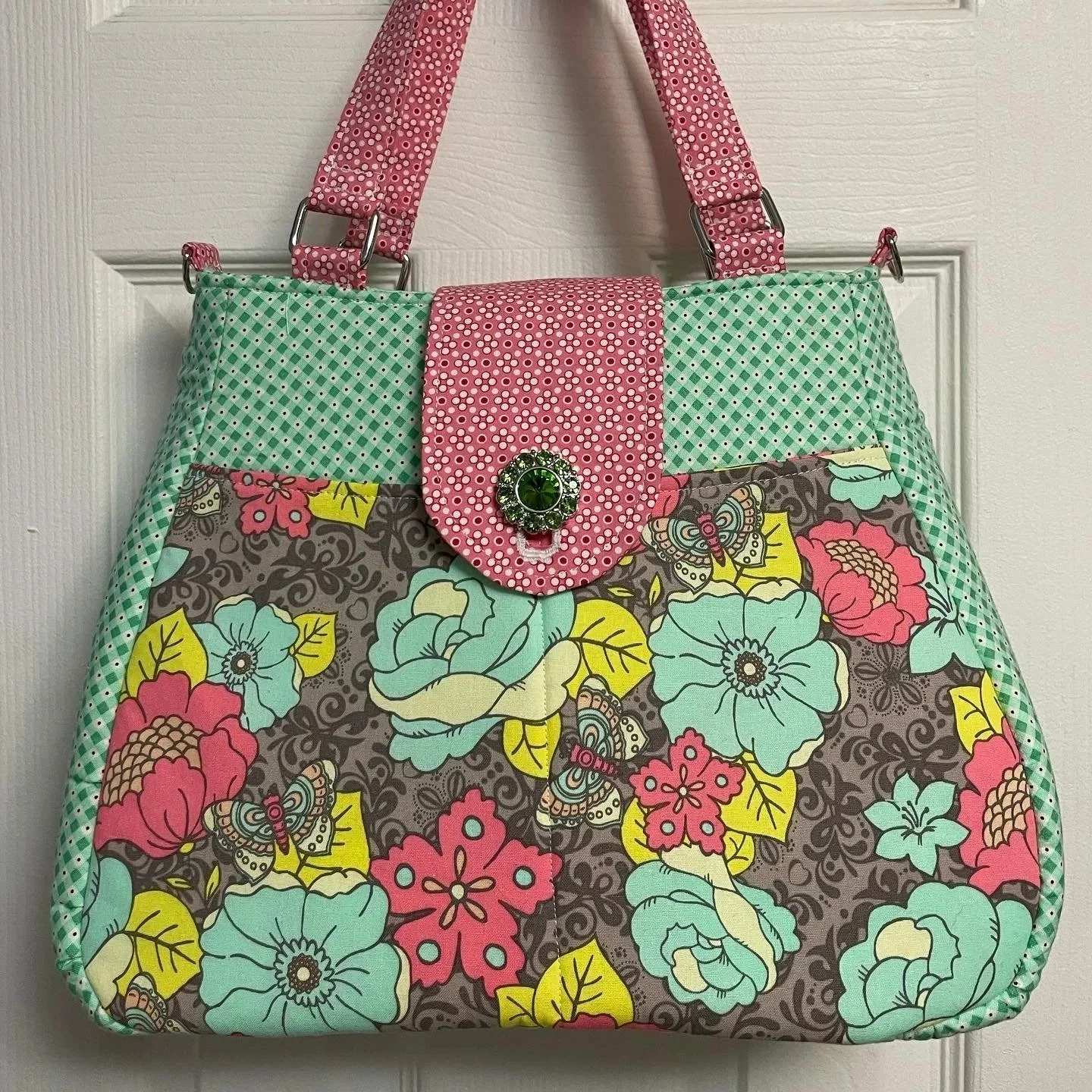 A colorful quilted tote bag with floral and butterfly patterns, pink dotted flap with a green gem button, and pink and green straps.