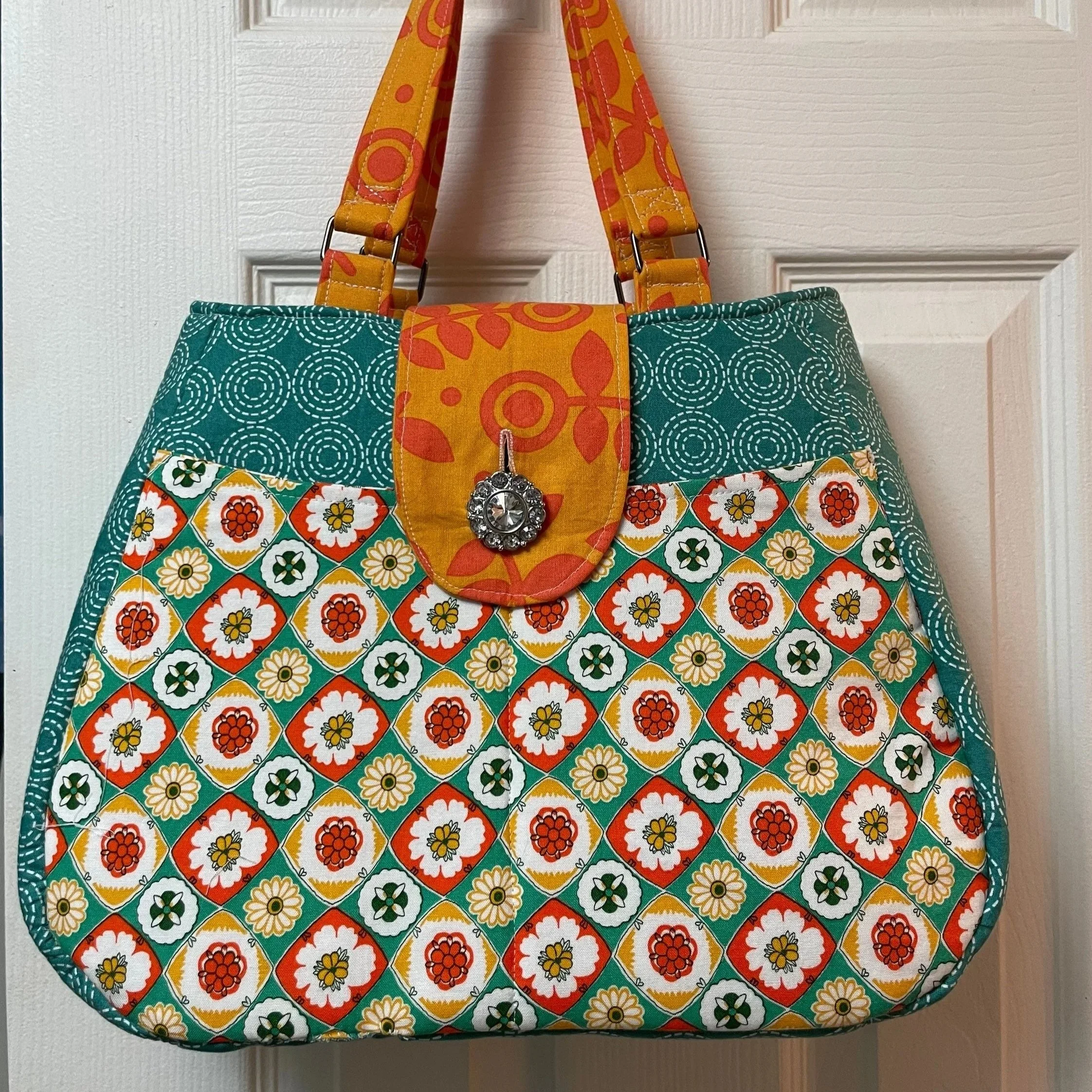 A colorful fabric tote bag hanging on a white door. The bag features a floral pattern with red, white, yellow, and green flowers, and contrasting teal and yellow sections with patterns. It has a decorative button on the flap.