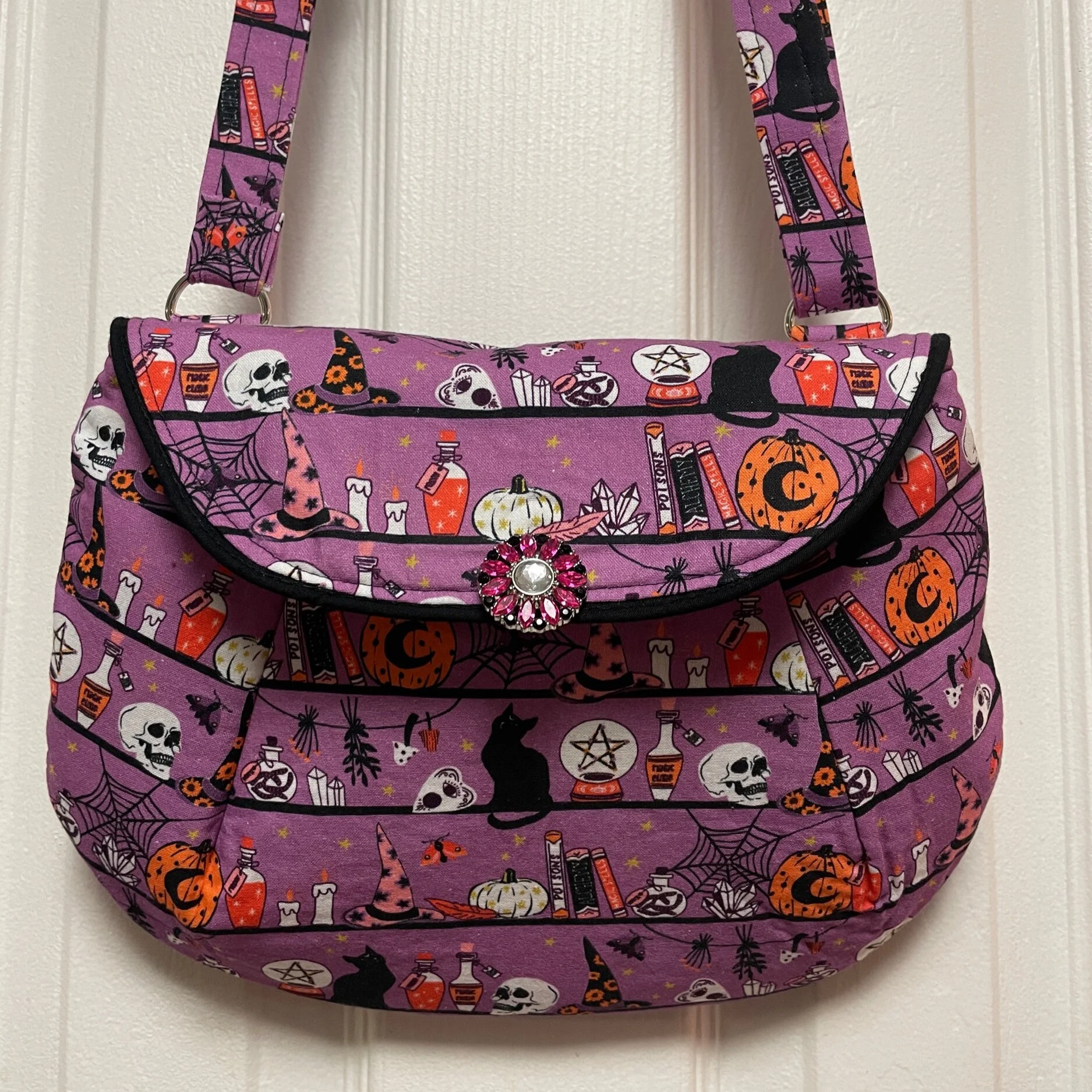 A purple Halloween-themed bag with illustrations of skulls, spiders, black cats, witches' hats, pumpkins, potion bottles, candles, owls, and books, featuring a decorative pink jewel on the flap.