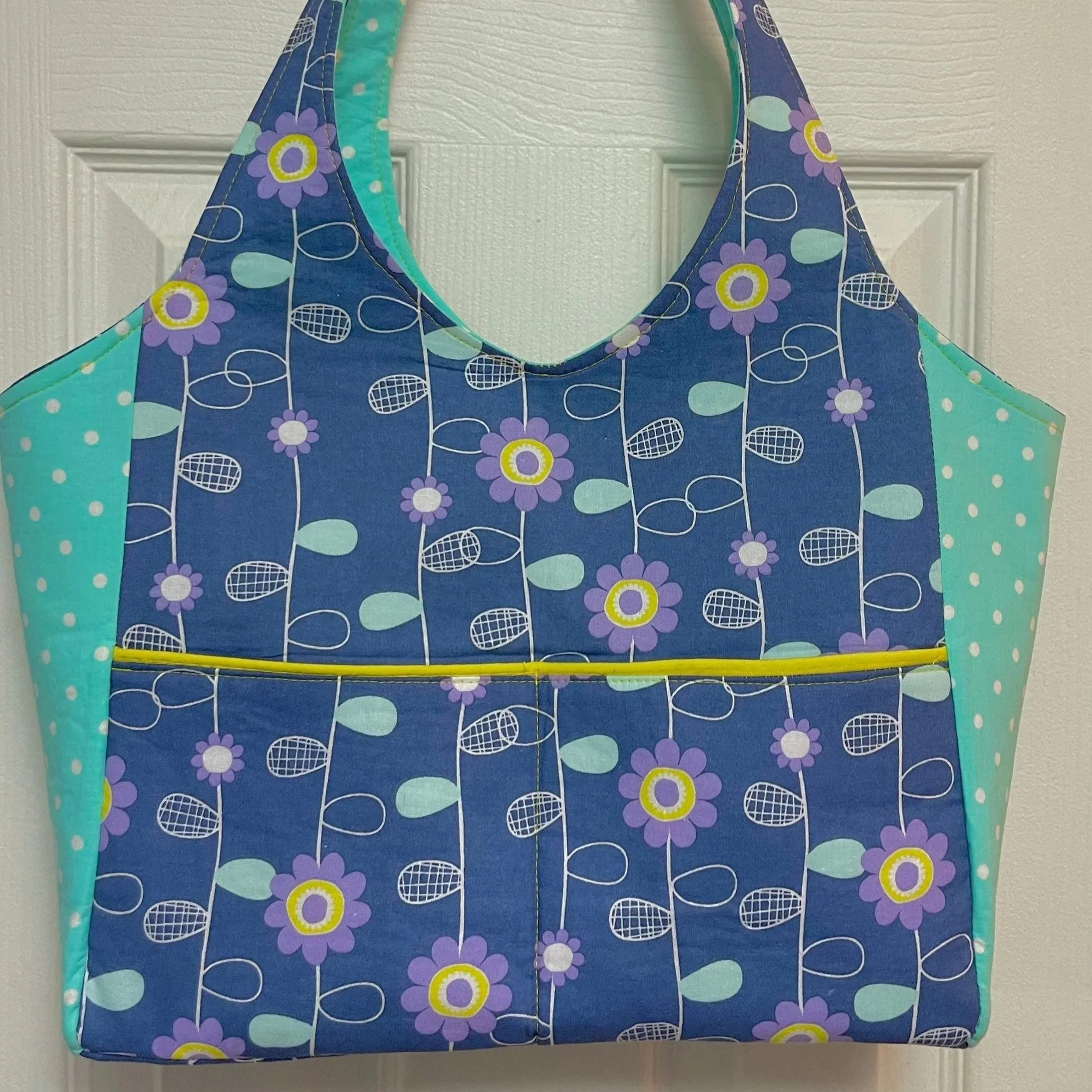 Bag with a blue floral pattern featuring purple and yellow flowers, and aqua polka dots on the sides, hanging on a white door.