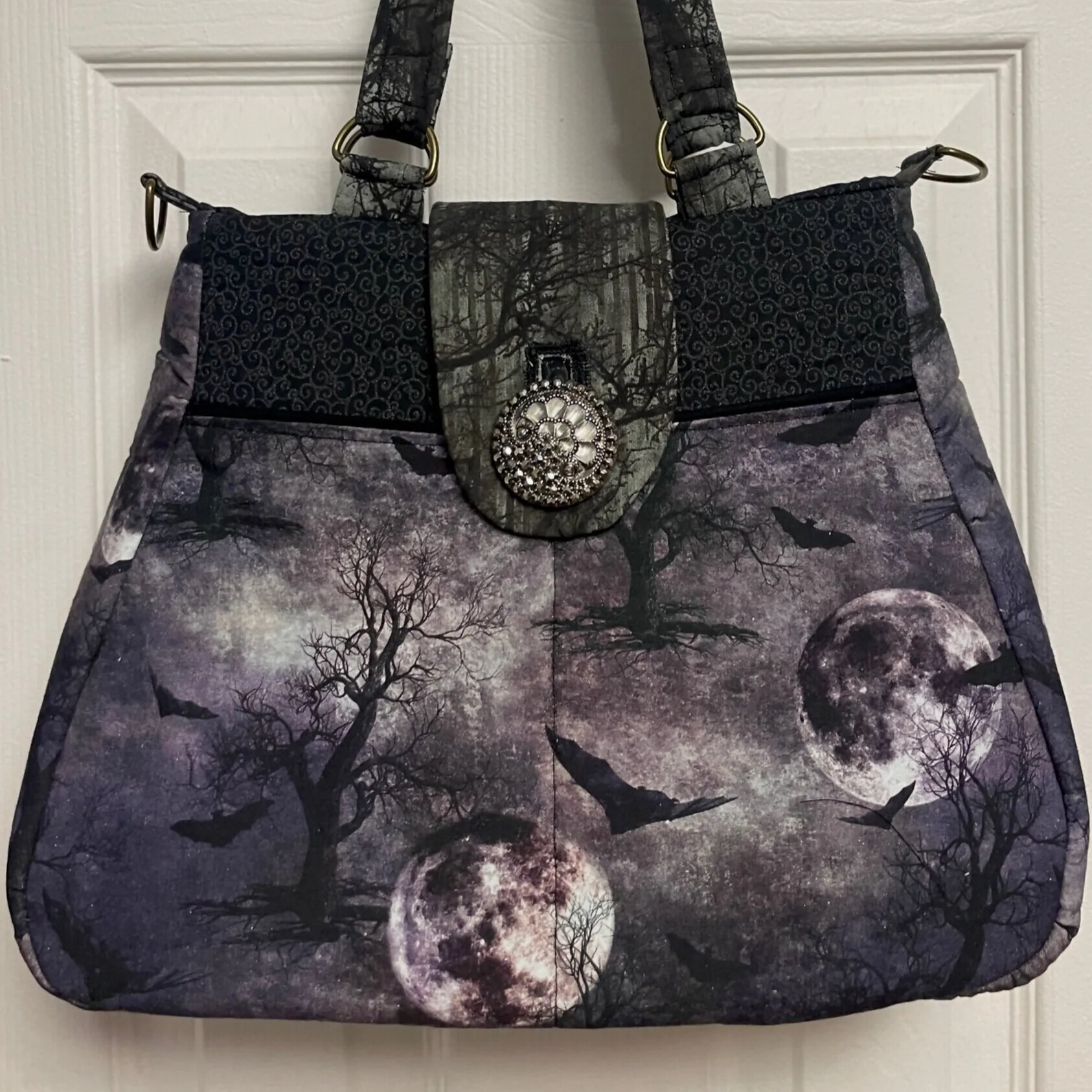 A Halloween-themed tote bag with a moonlit forest and bats design, decorated with a black and silver brooch.
