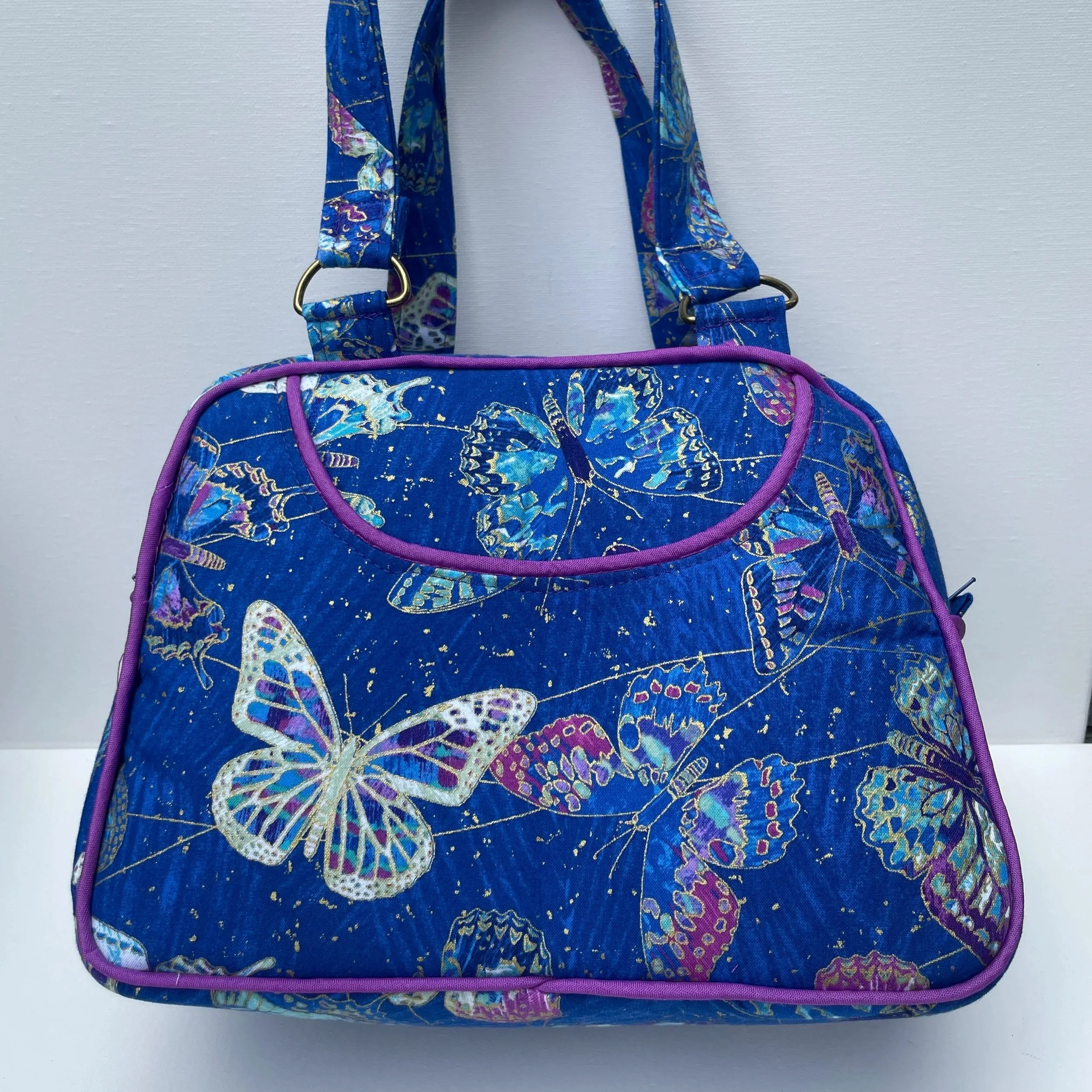 A blue fabric bag with a butterfly pattern and purple piping, hanging against a white wall.