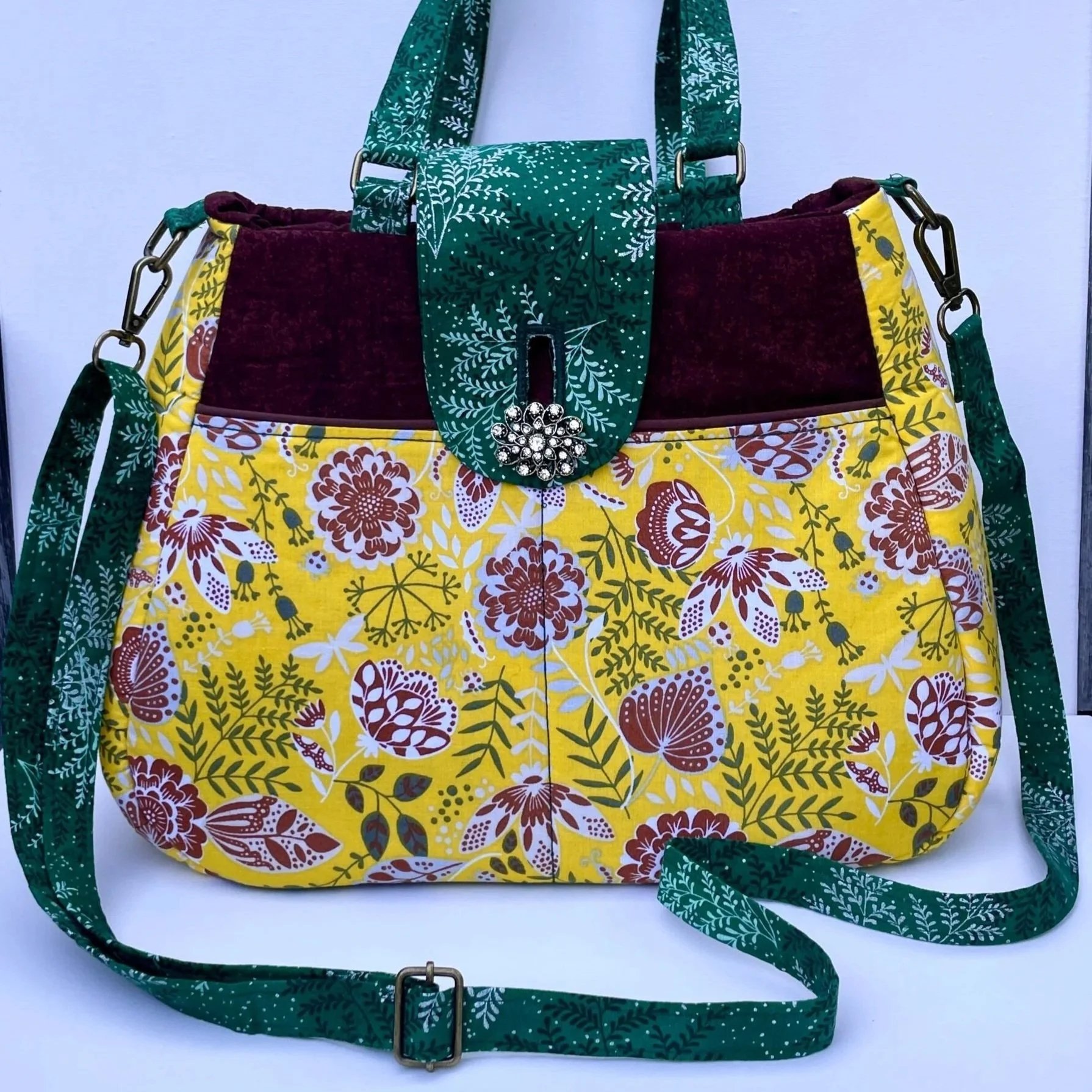 A colorful tote bag with a yellow floral pattern, maroon and green accents, and a green strap with white leaf designs.