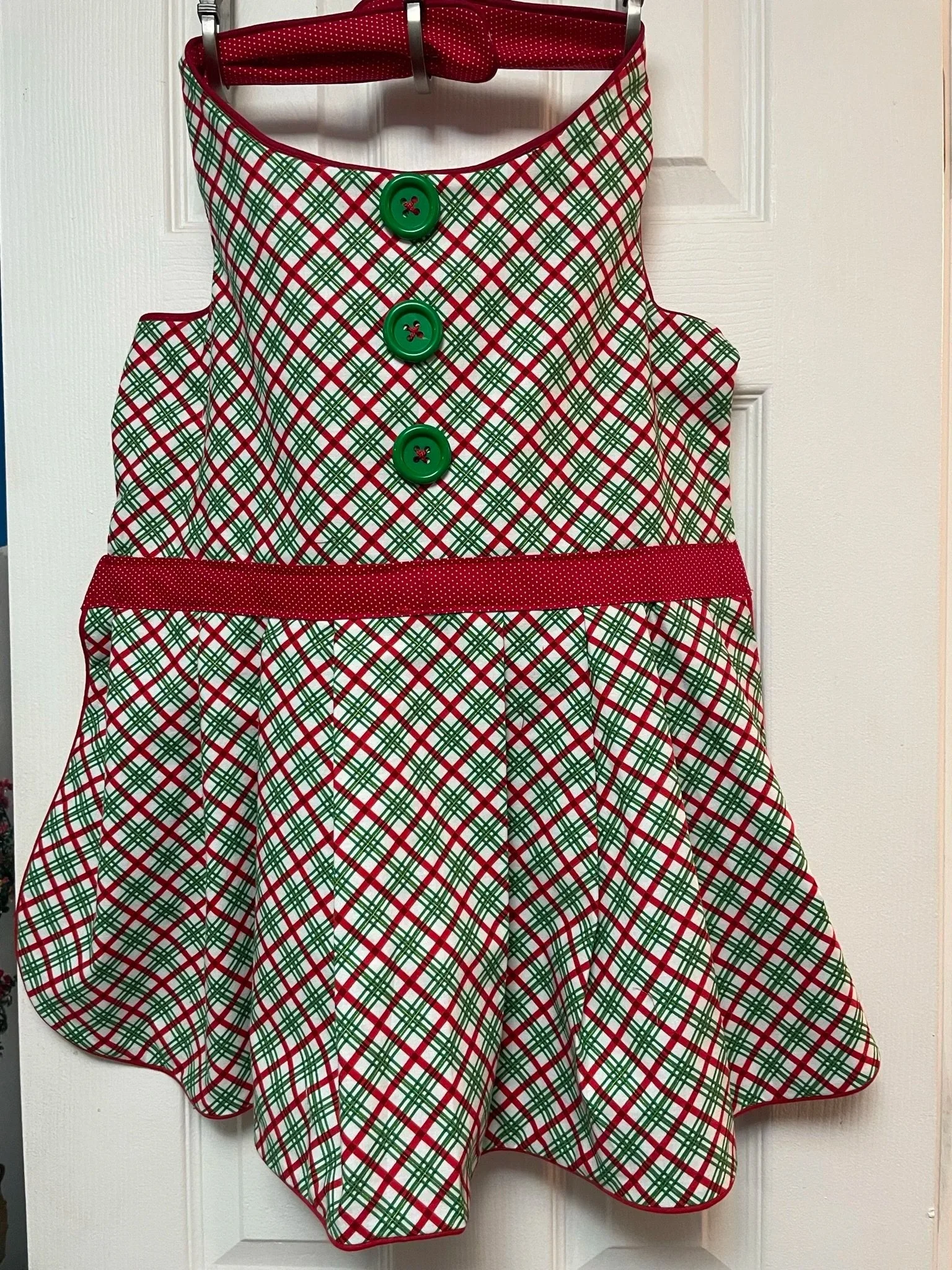 A festive sleeveless dress with a red, green, and white plaid pattern, four large green buttons down the front, a red waistband, and red trim at the neckline and hem, hanging on a hanger in front of a white door.