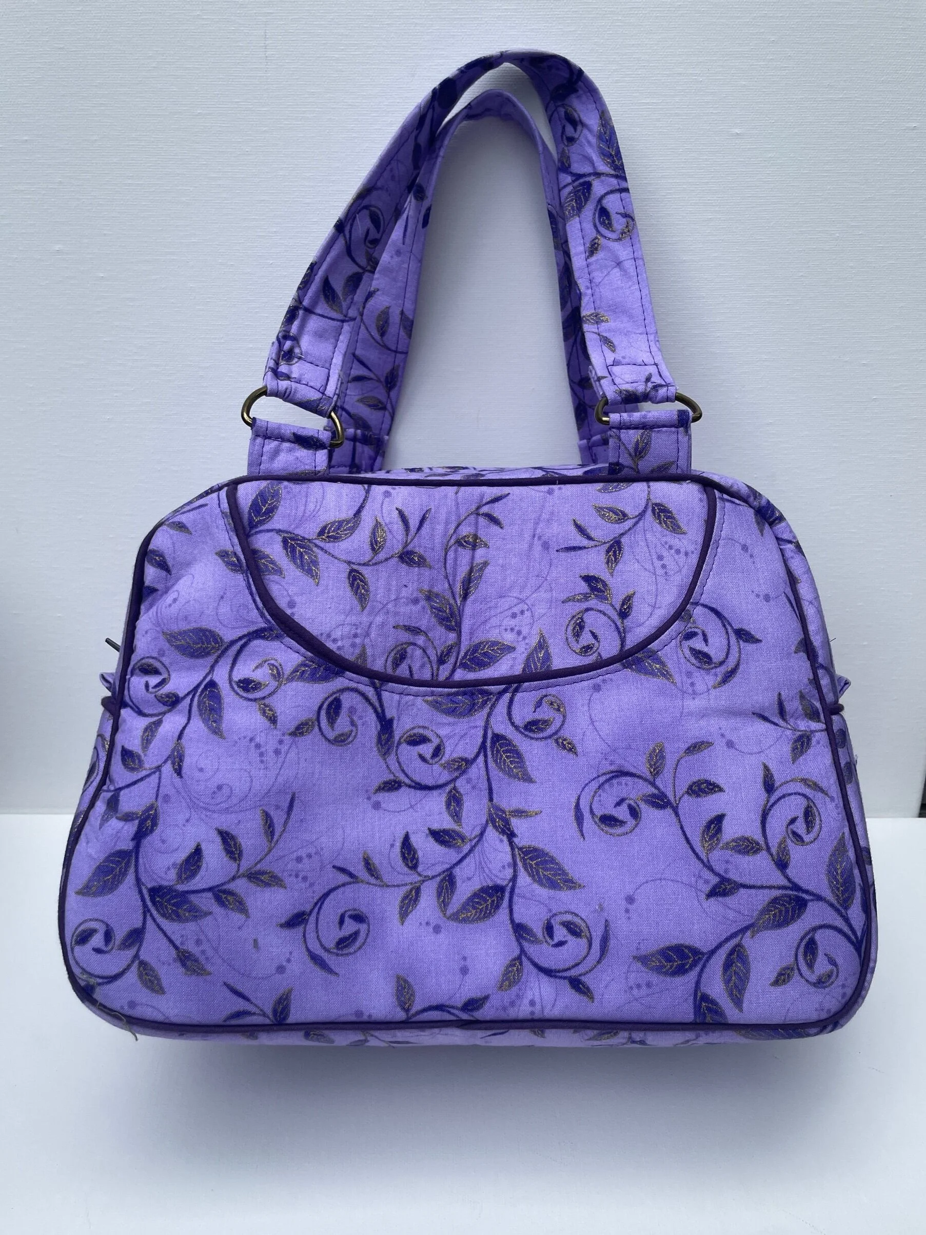 A purple handbag with a floral pattern and two handles, hanging on a white wall.