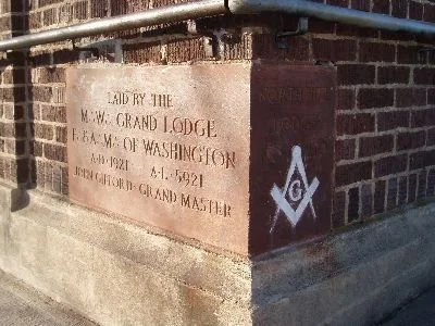 A stone monument with engraved text about the Masonic Grand Lodge of Washington, including a Masonic square and compass symbol painted on the side of a brick building.