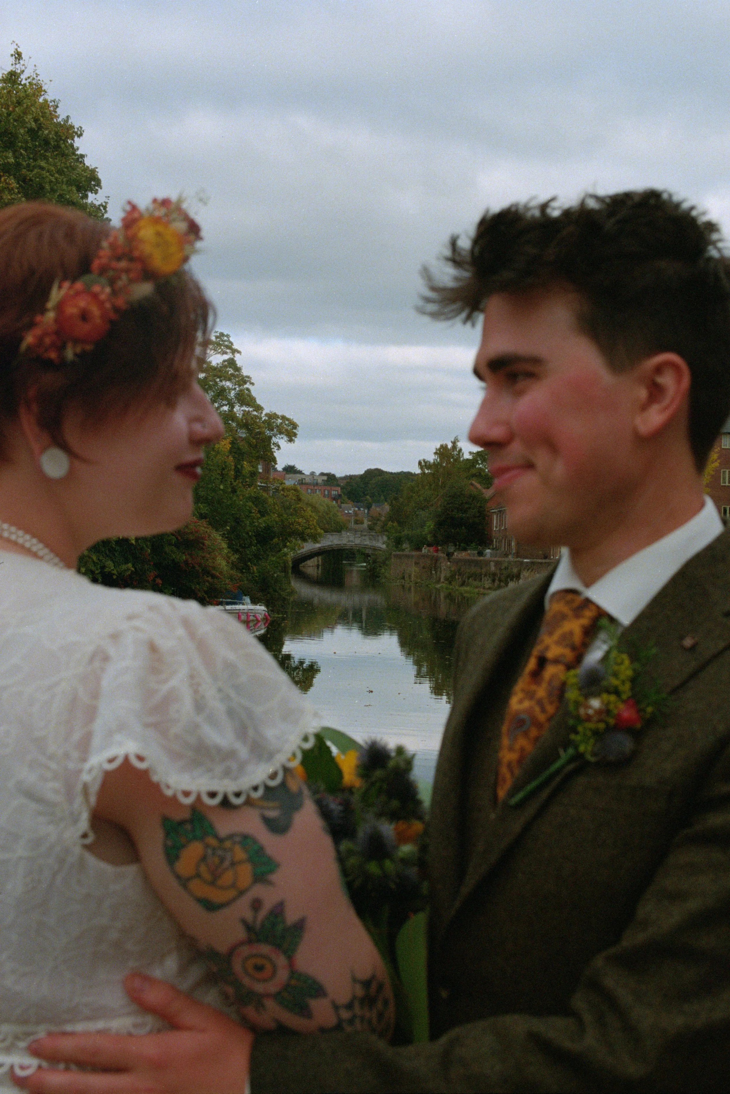 A bride and groom stand close together by a river, gazing into each other's eyes, with a bridge and trees in the background.