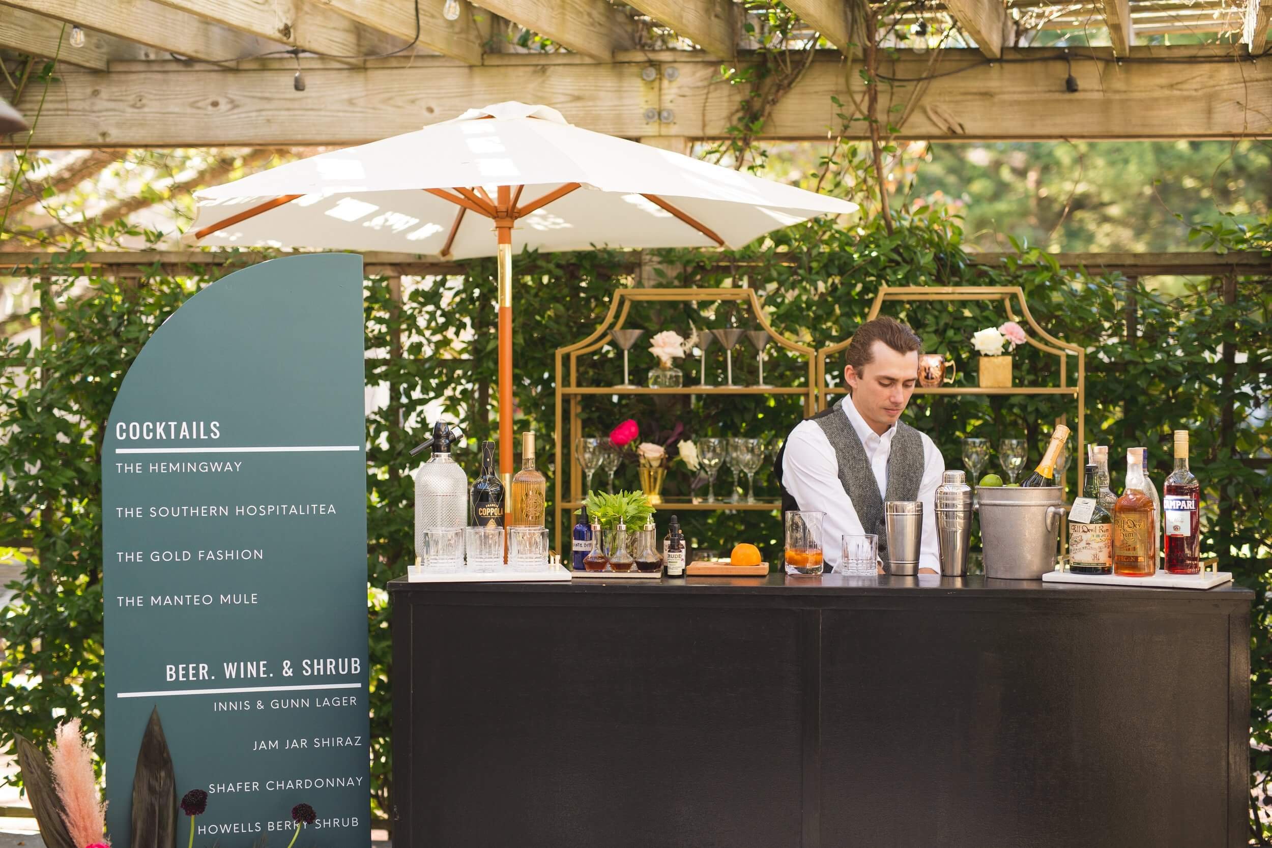  A man standing behind a bar with custom decor rentals and umbrella at Outer Banks wedding 