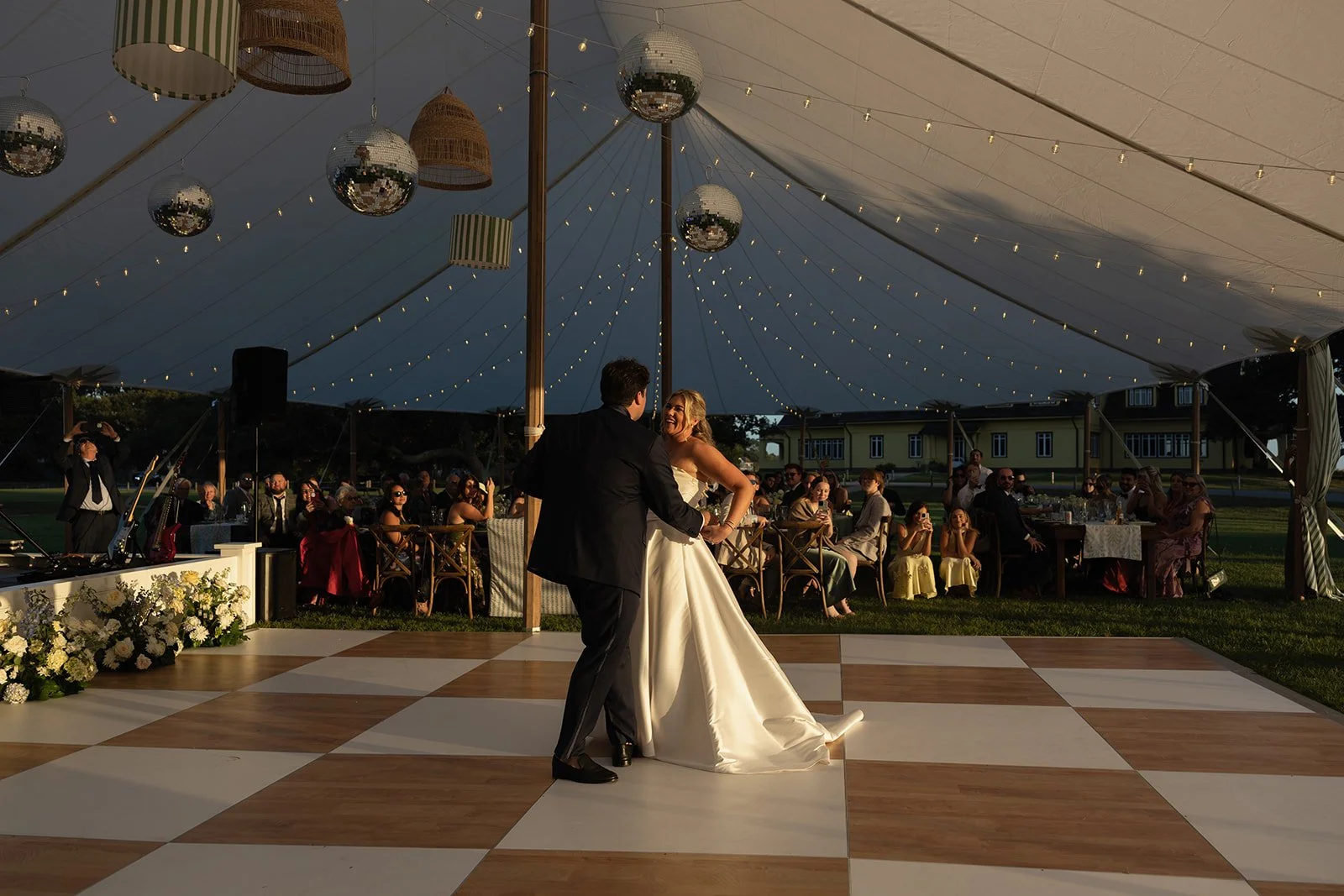 Bride and groom dancing on a checkered dance floor beneath disco balls at a tented wedding reception, styled with designer event rentals by Amaranth + Artifact