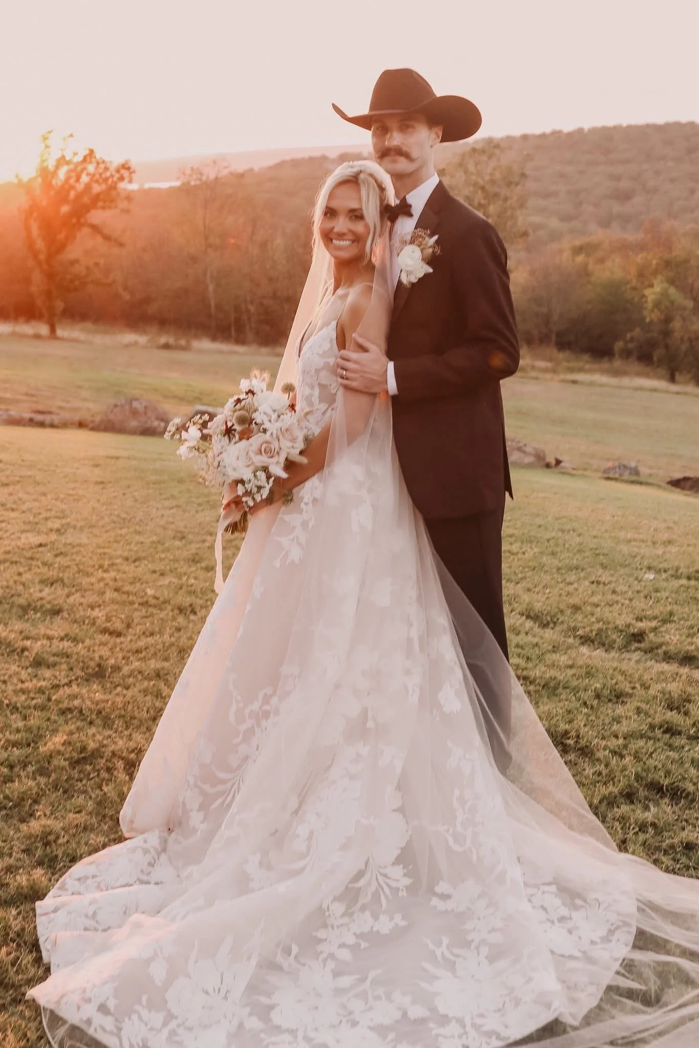 Bride and groom in wedding attire sharing a romantic moment outdoors, surrounded by breathtaking sunset. The bride holds a bouquet of fall inspired flowers and wears a designer wedding gown.