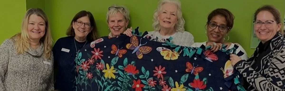 Six women standing together indoors, holding a colorful floral and butterfly blanket, in front of lime green wall.