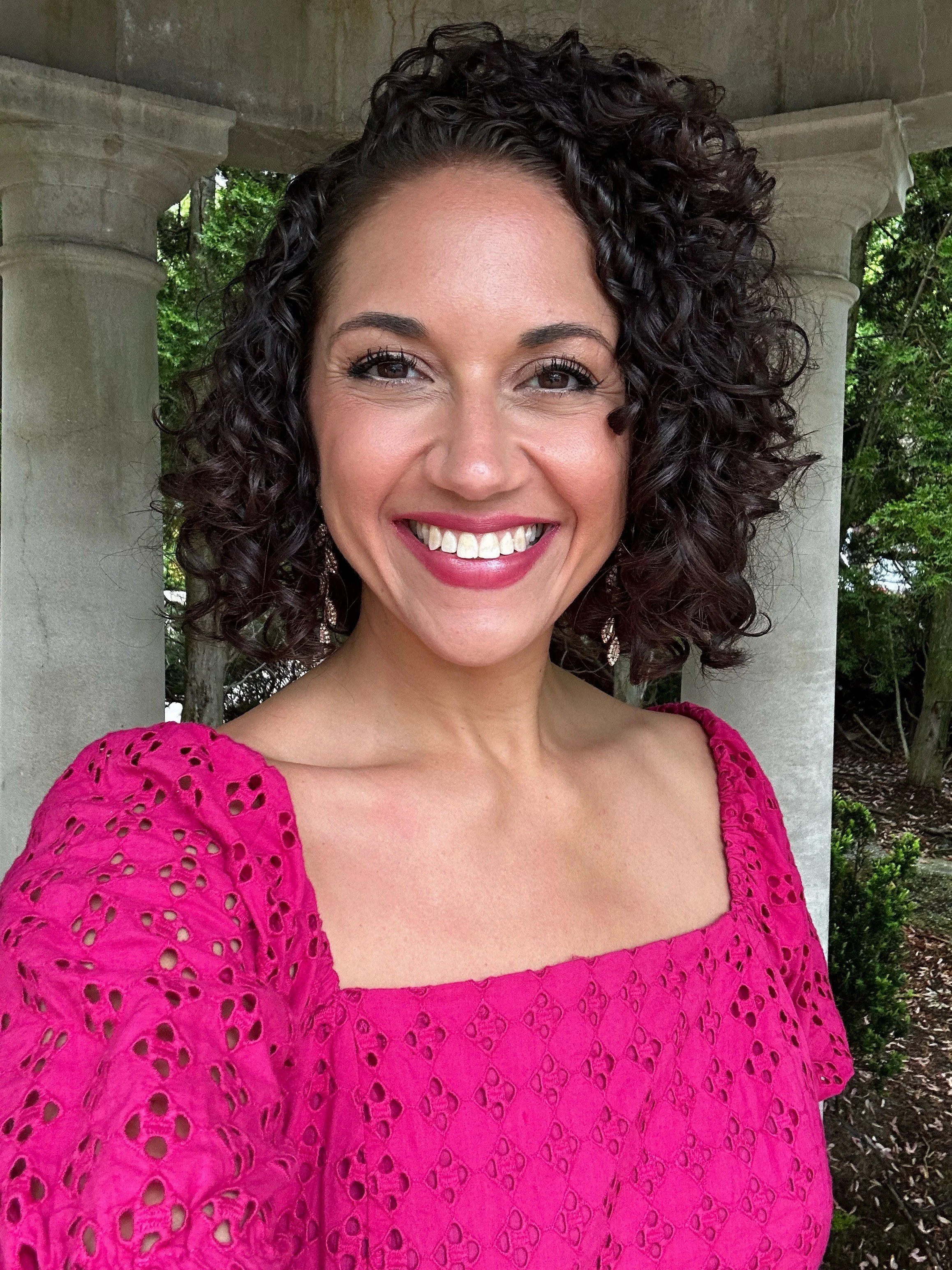 A woman with curly dark hair smiling, wearing a pink eyelet dress and earrings.