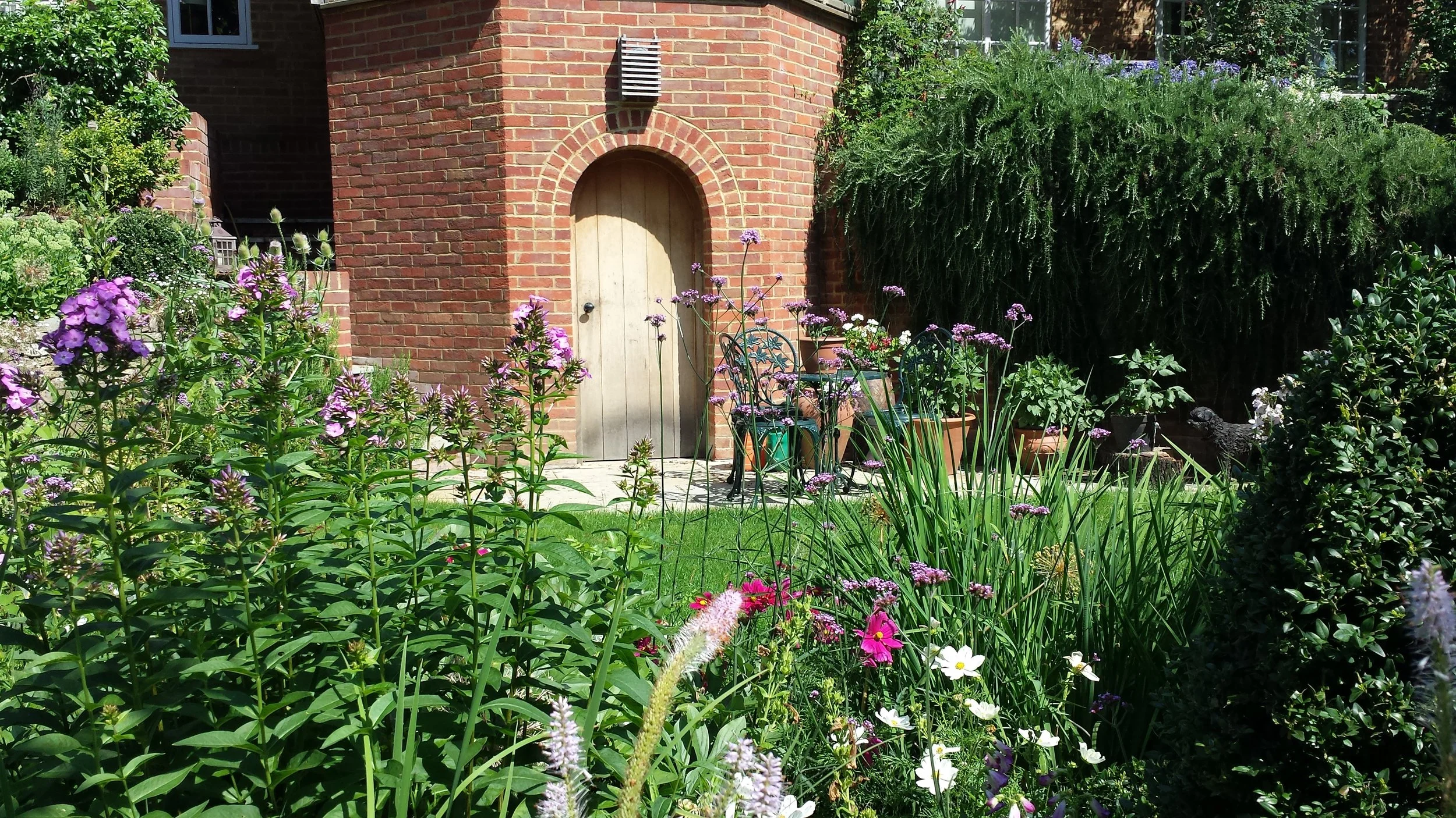 A lush garden in front of a brick house with a rounded wooden door, colorful flowers, and green foliage, with outdoor chairs and potted plants.