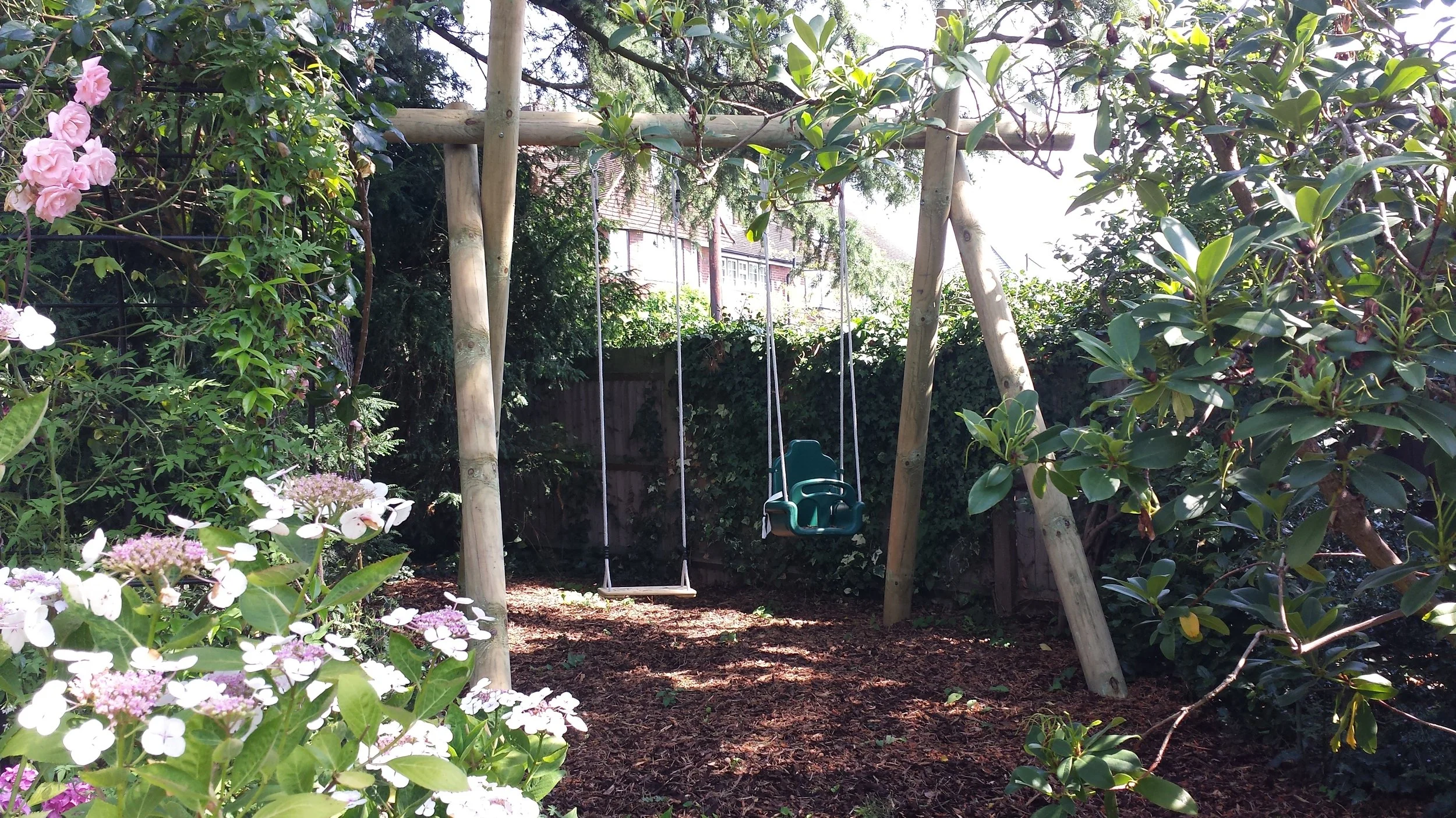 A backyard garden with a wooden swing set and green toddler swing, surrounded by lush garden plants and pink and white flowers, with trees and houses in the background.