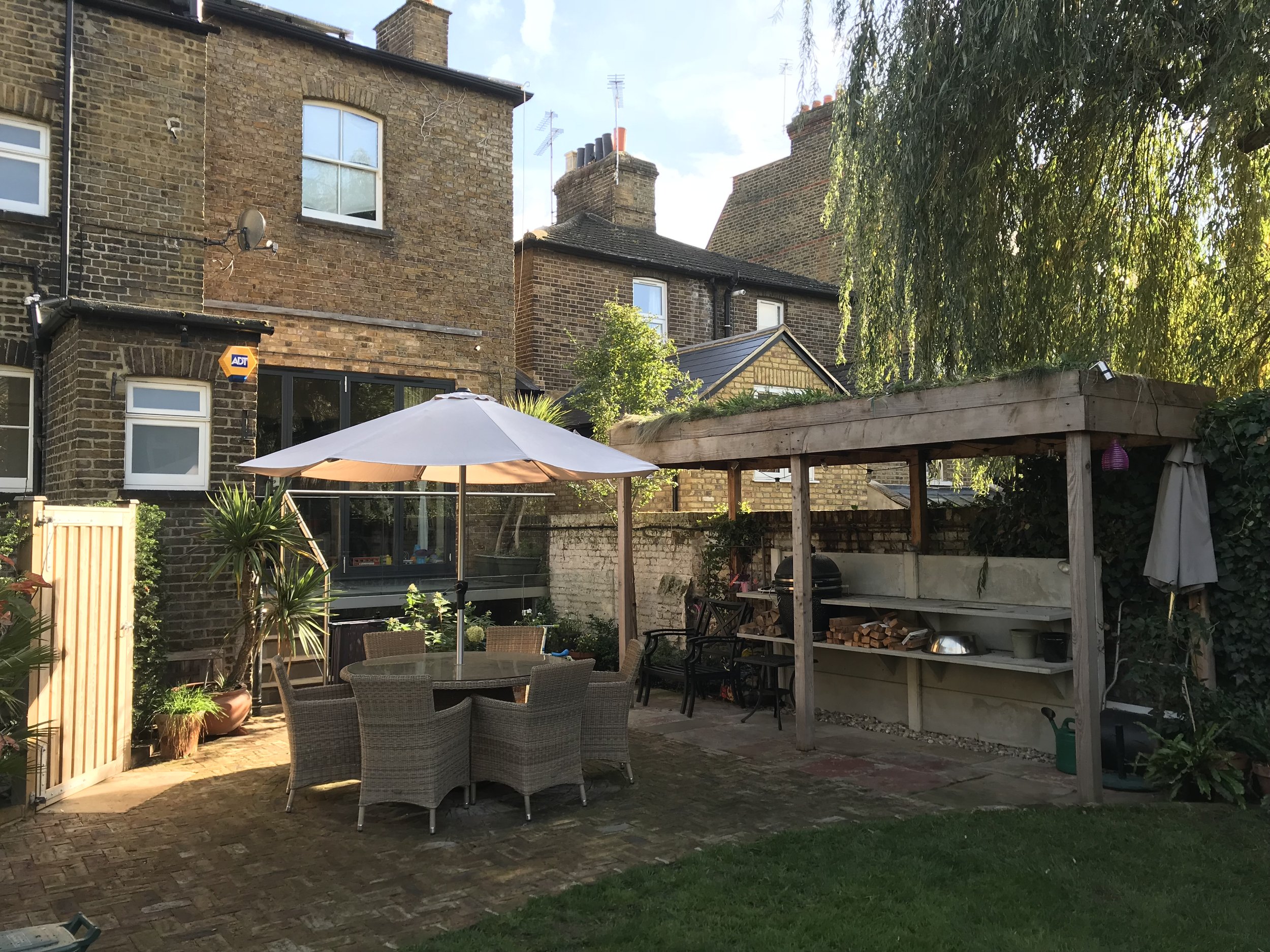 Backyard patio with round table and six chairs, large white umbrella, wooden shed with shelves, brick paving, potted plants, and a lush green lawn.