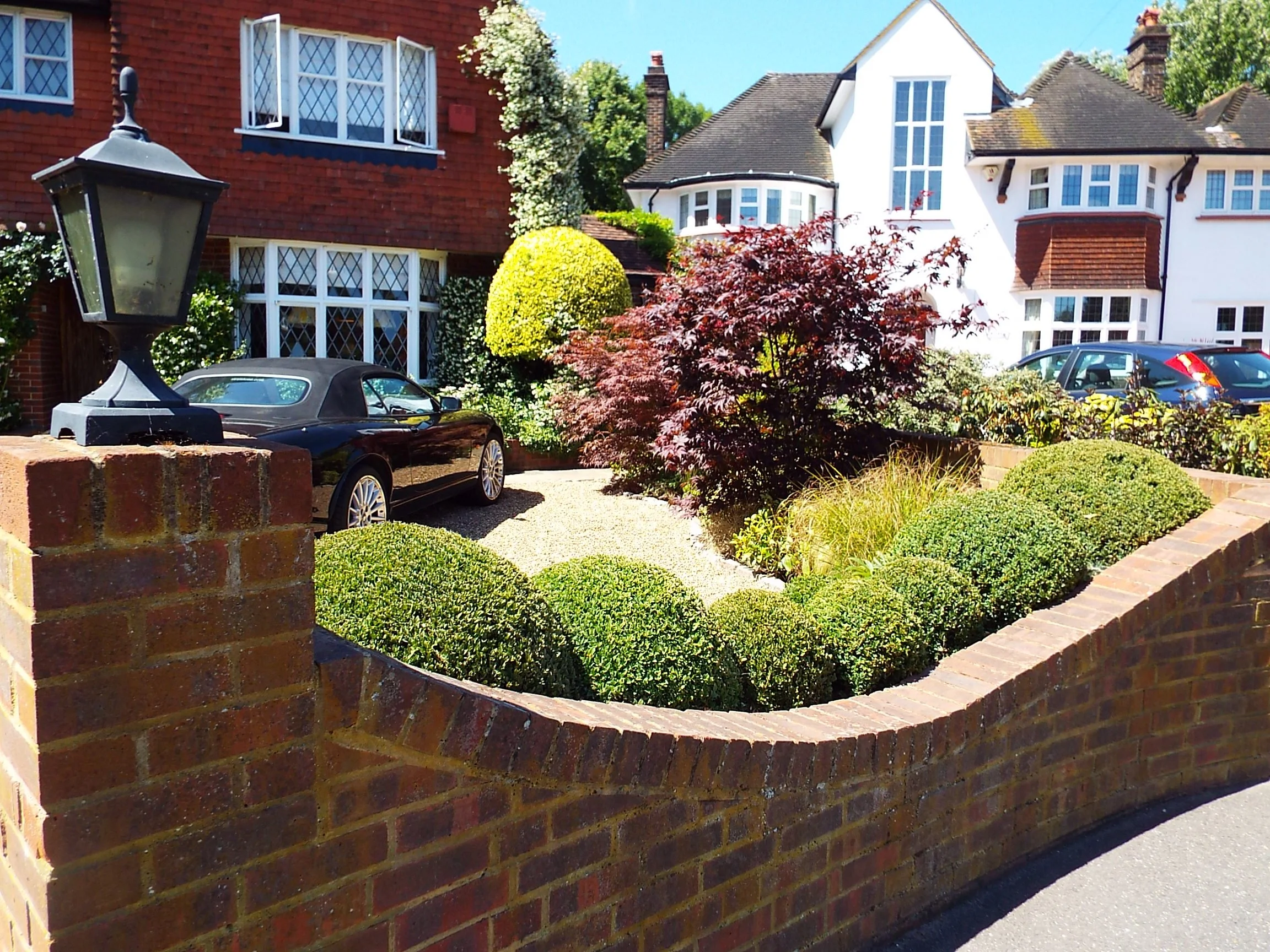A brick wall with a lamp post on it surrounds a landscaped front yard with bushes, a small tree with red leaves, a black convertible car, and a gravel driveway in front of residential houses with white and red facades.