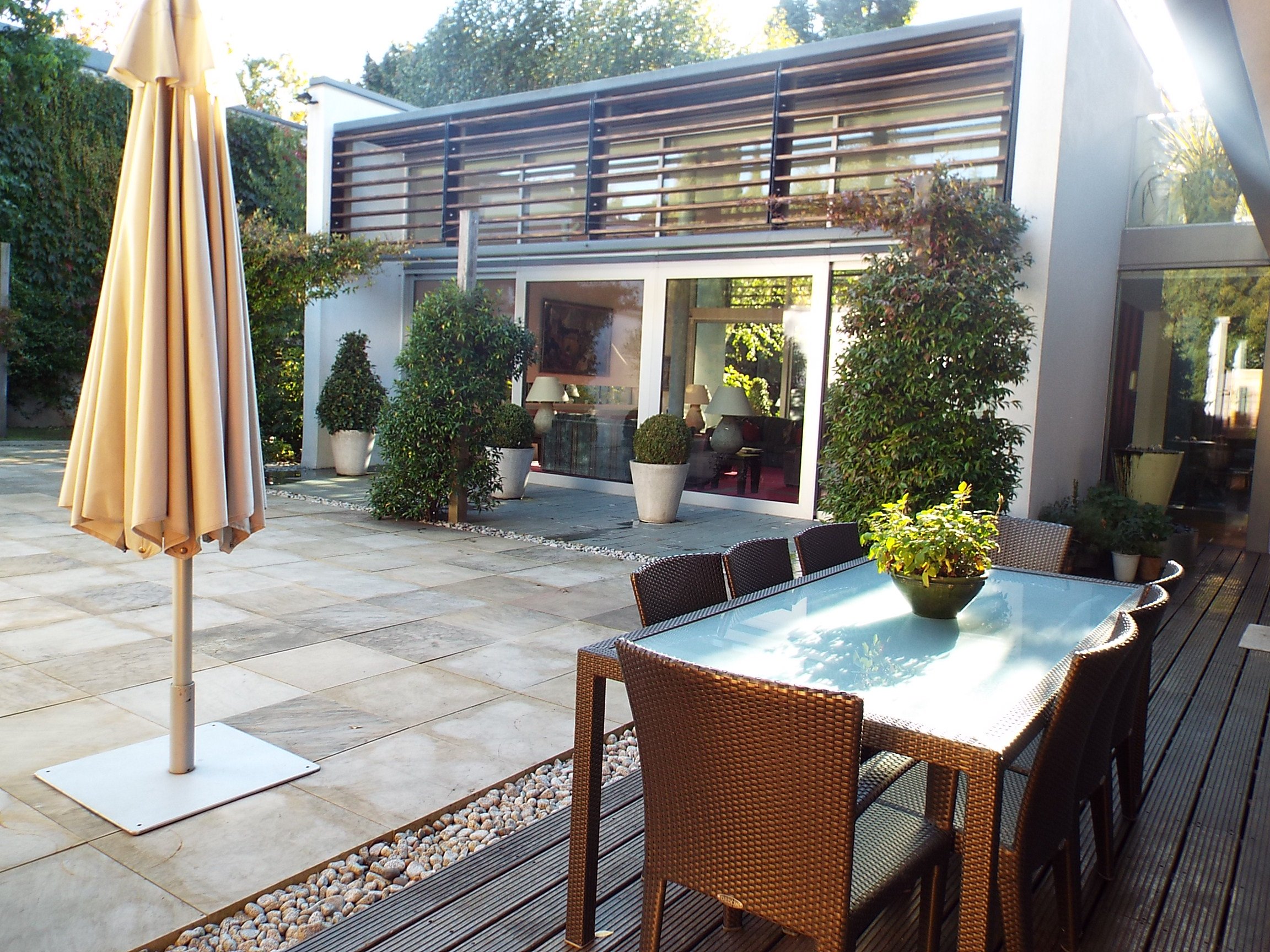 Outdoor patio featuring a glass-topped dining table with six chairs, potted plants, a closed beige umbrella, and a modern house with large glass windows and green plants in the background.