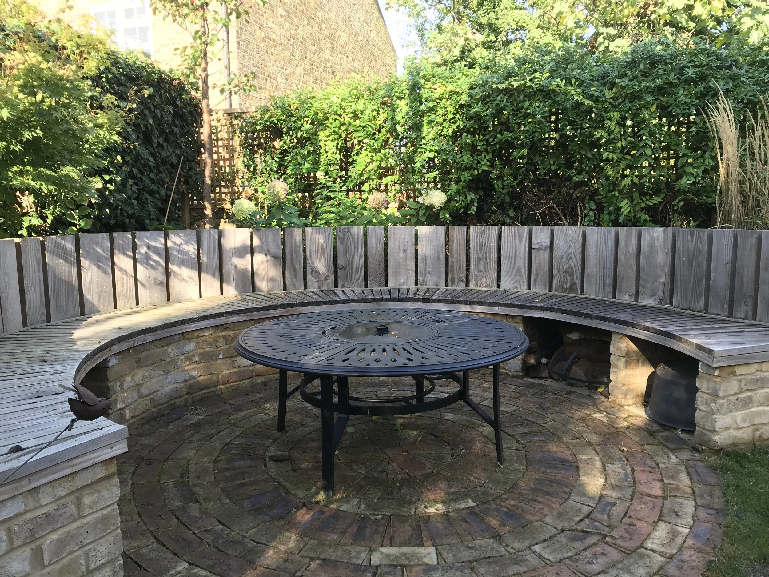 A semi-circular wooden bench surrounds a round metal fire pit on a brick patio with green shrubs and a brick wall in the background.