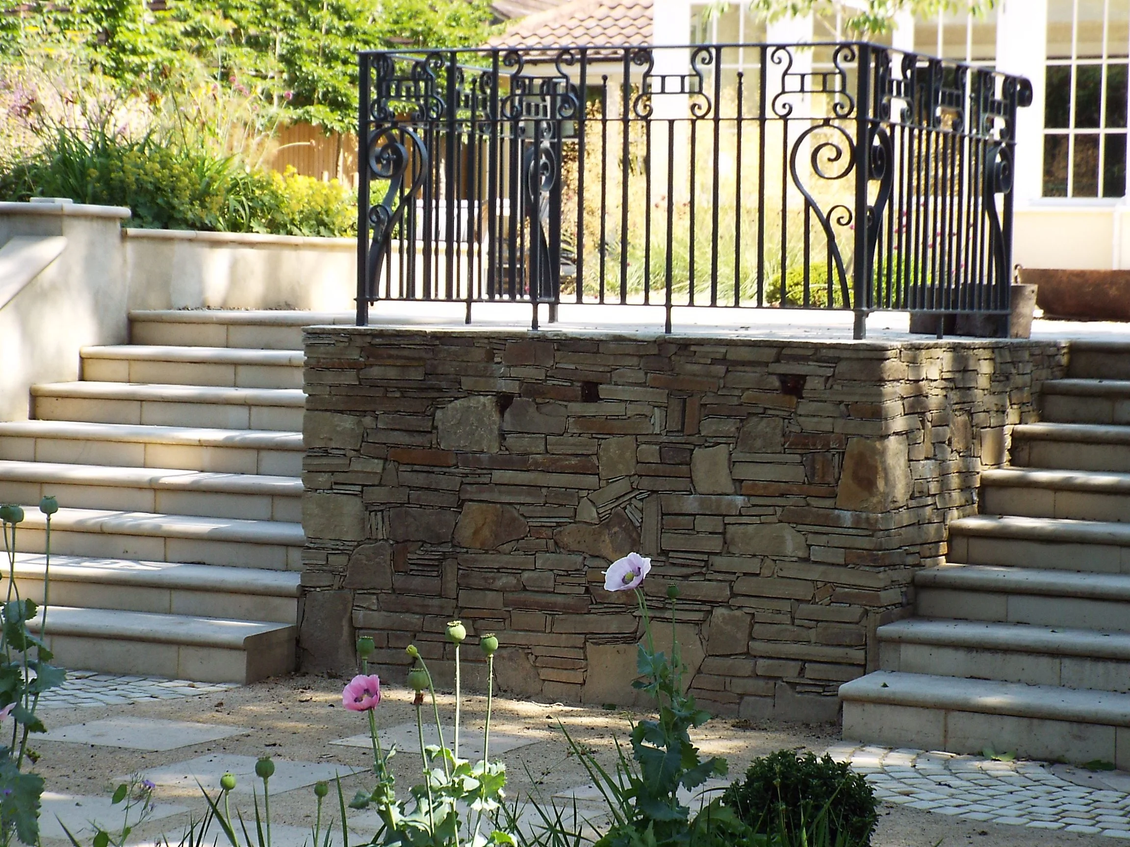 Outdoor stone and tile patio with stairs, black iron railing, and flower garden.
