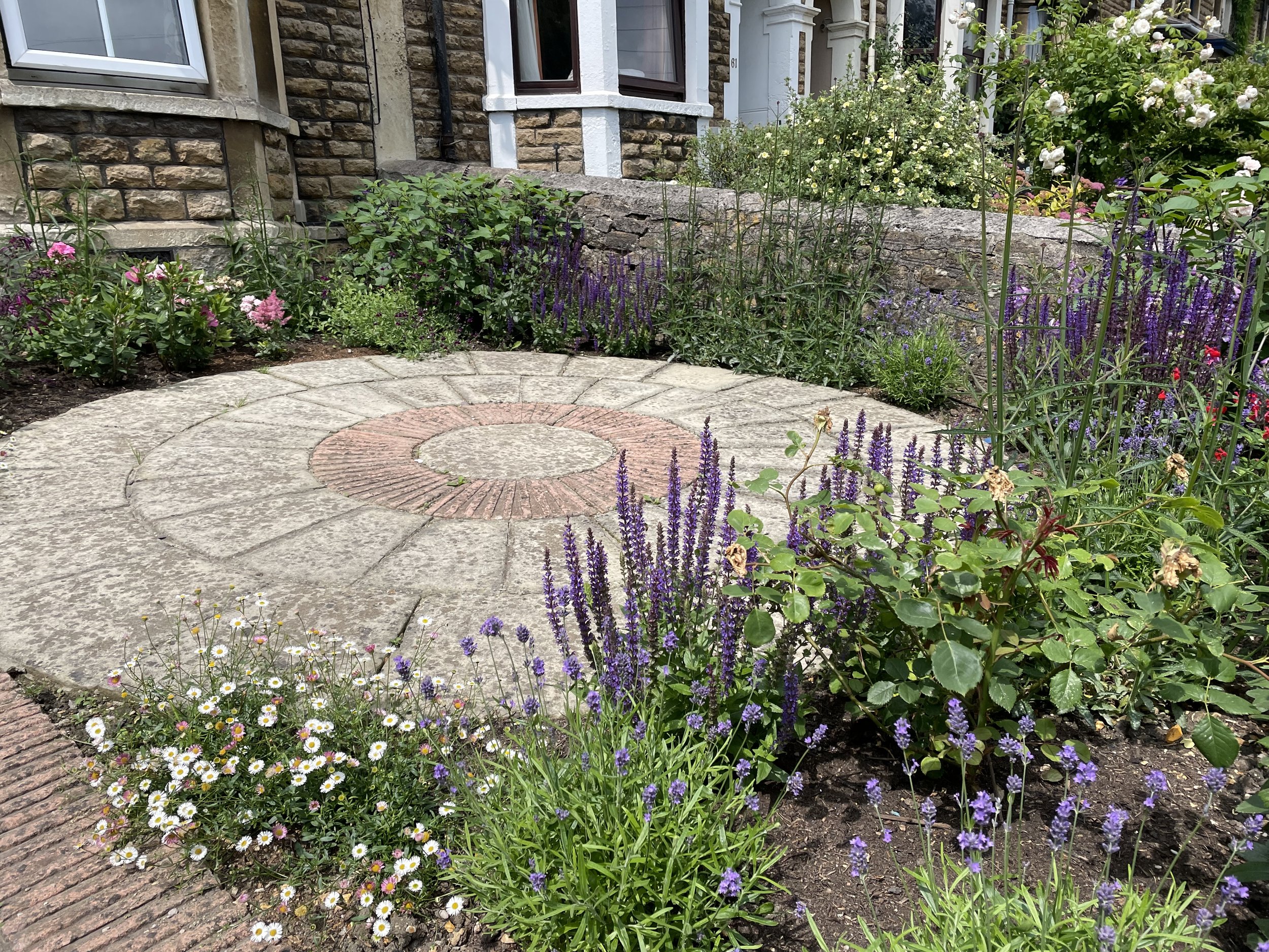 A circular stone patio surrounded by colorful flowering plants and bushes in front of a house with a brick and white painted exterior.