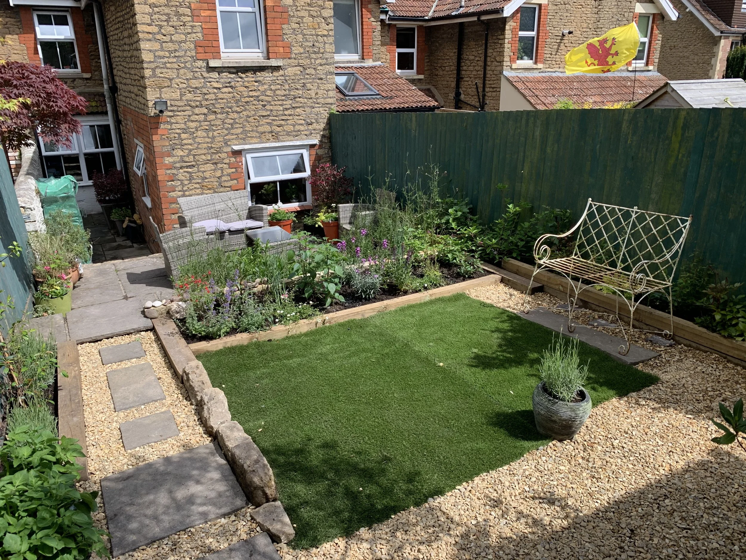 A small backyard garden with a green fence, a patch of artificial grass, a potted lavender plant, and a white wrought iron bench. There are flowering plants along the border, a stone pathway, and outdoor seating with cushions against the house.
