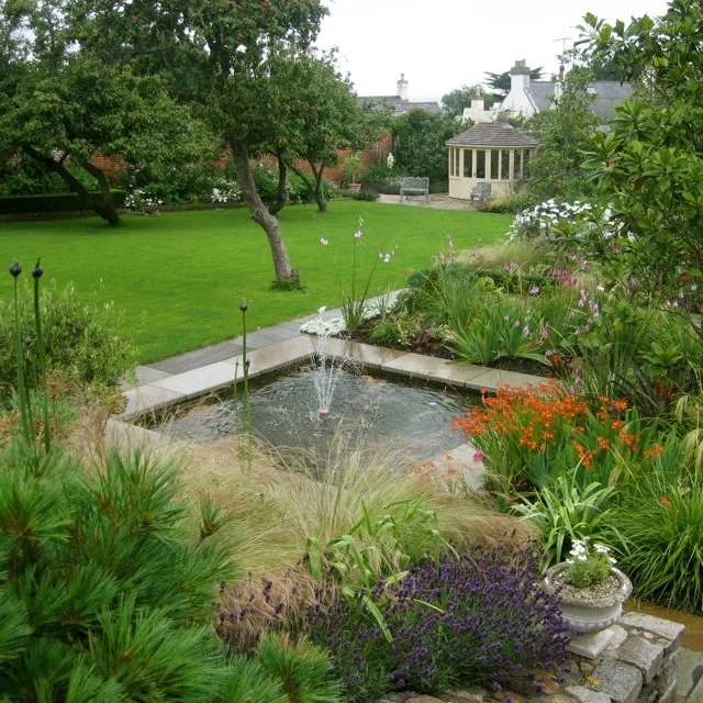 A lush garden with a small fountain, surrounded by various green plants and colorful flowers, with a grassy lawn and trees in the background.