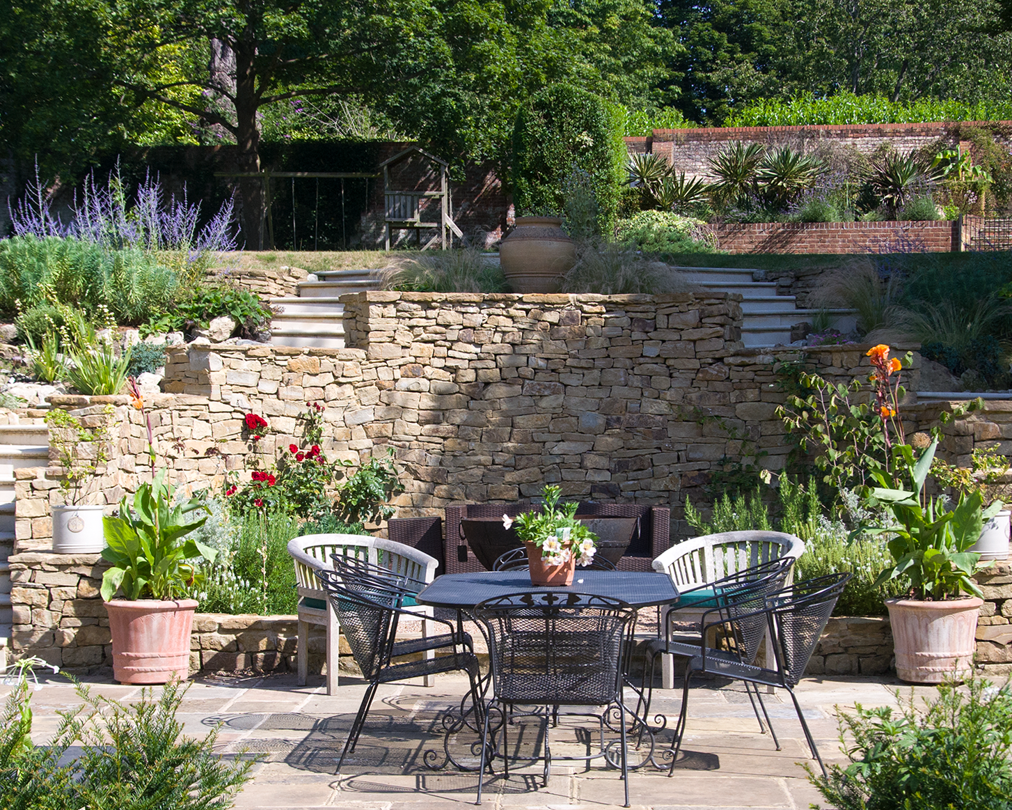 Outside garden patio with black metal dining table and chairs, surrounded by potted plants and colorful flowers, stone terraces, lush green trees and plants in the background.