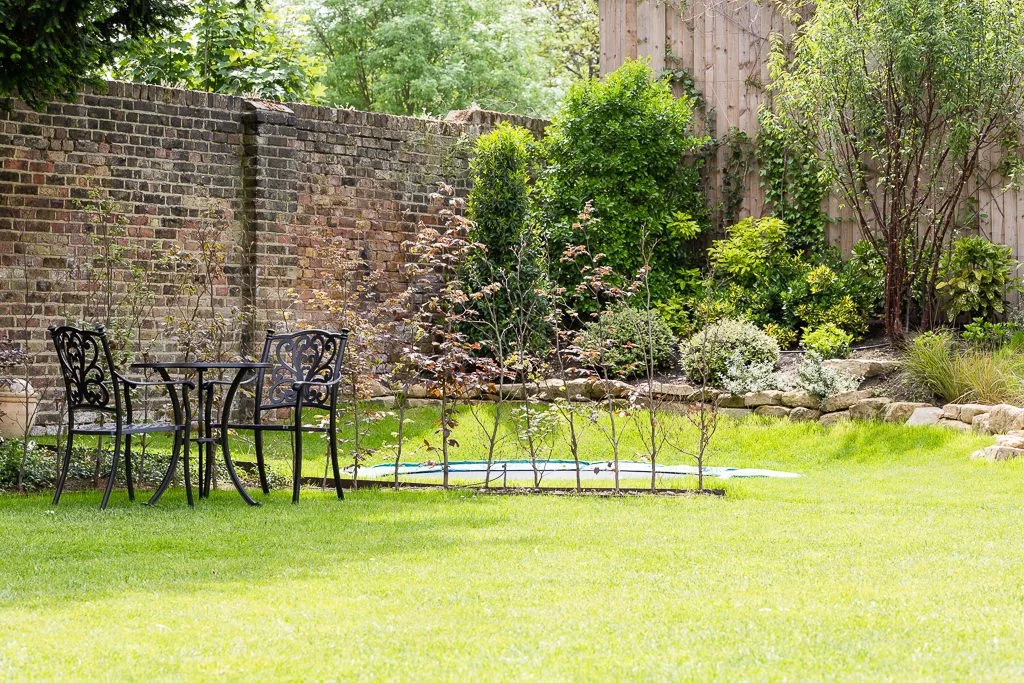 A backyard garden with a brick wall, green grass, small trees, shrubs, and a black metal table with two chairs.