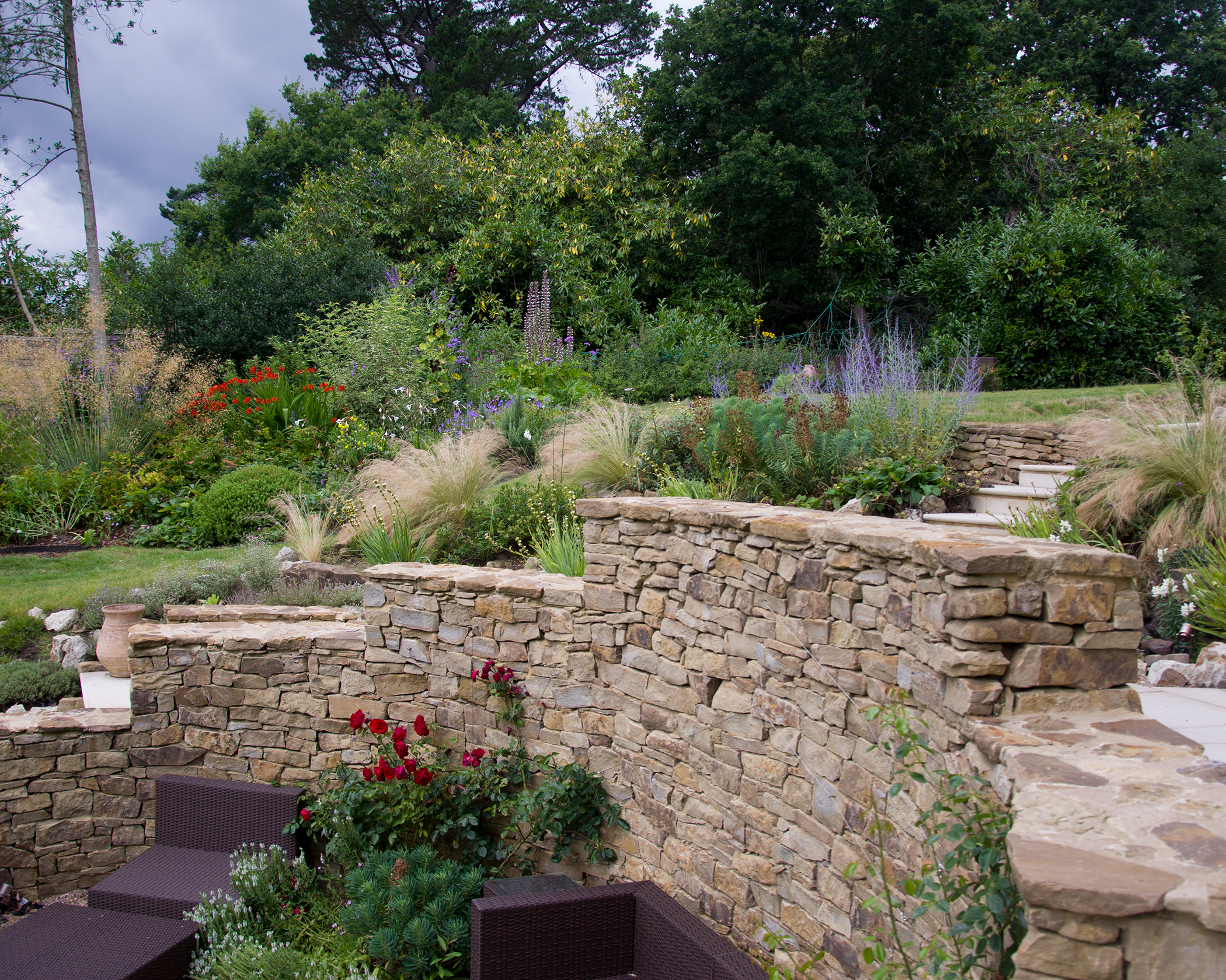 A backyard garden with stone retaining walls, a variety of flowering plants, lush greenery, and outdoor furniture.