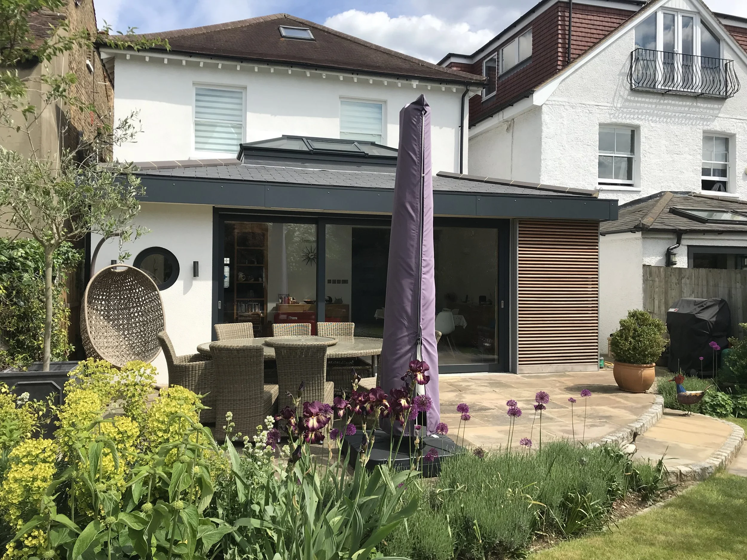 Backyard patio with wicker dining table and chairs, purple patio umbrella, potted plants, and bright garden flowers, with a modern house in the background.