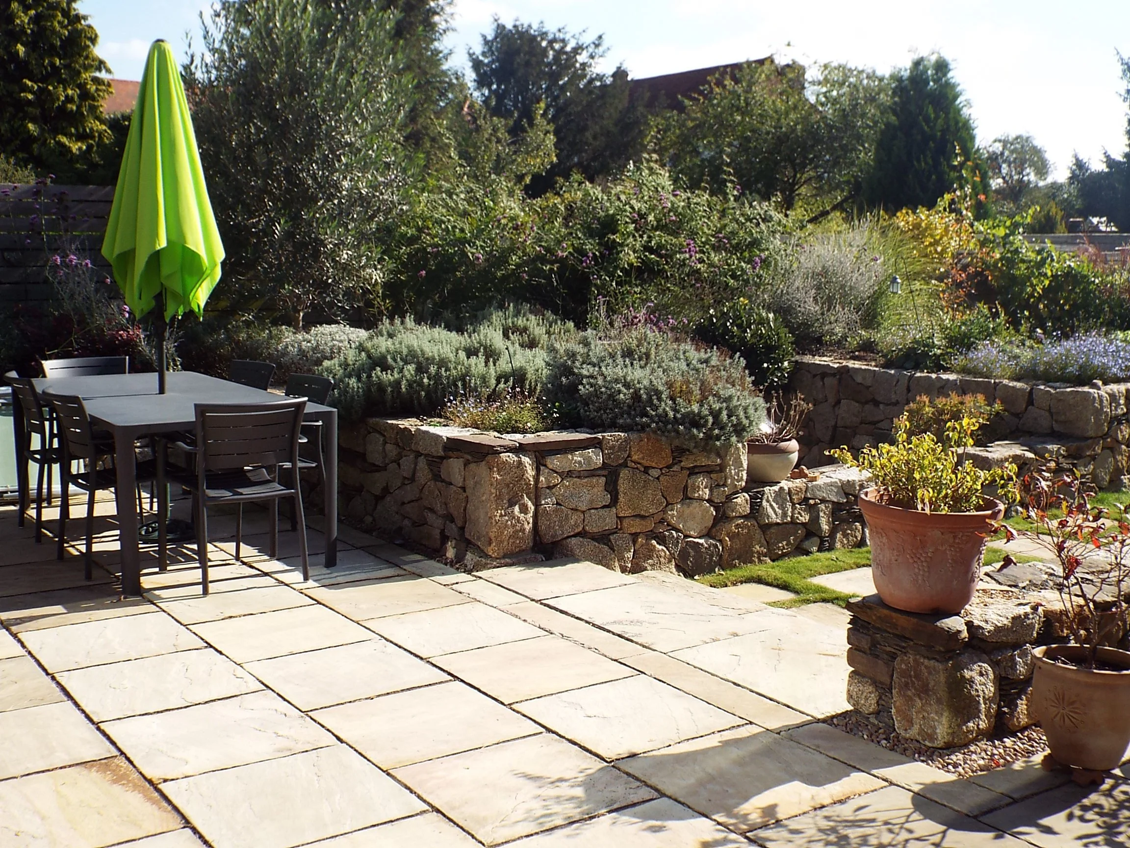 Patio with outdoor furniture, potted plants, stone retaining wall, and lush garden greenery in sunlight.