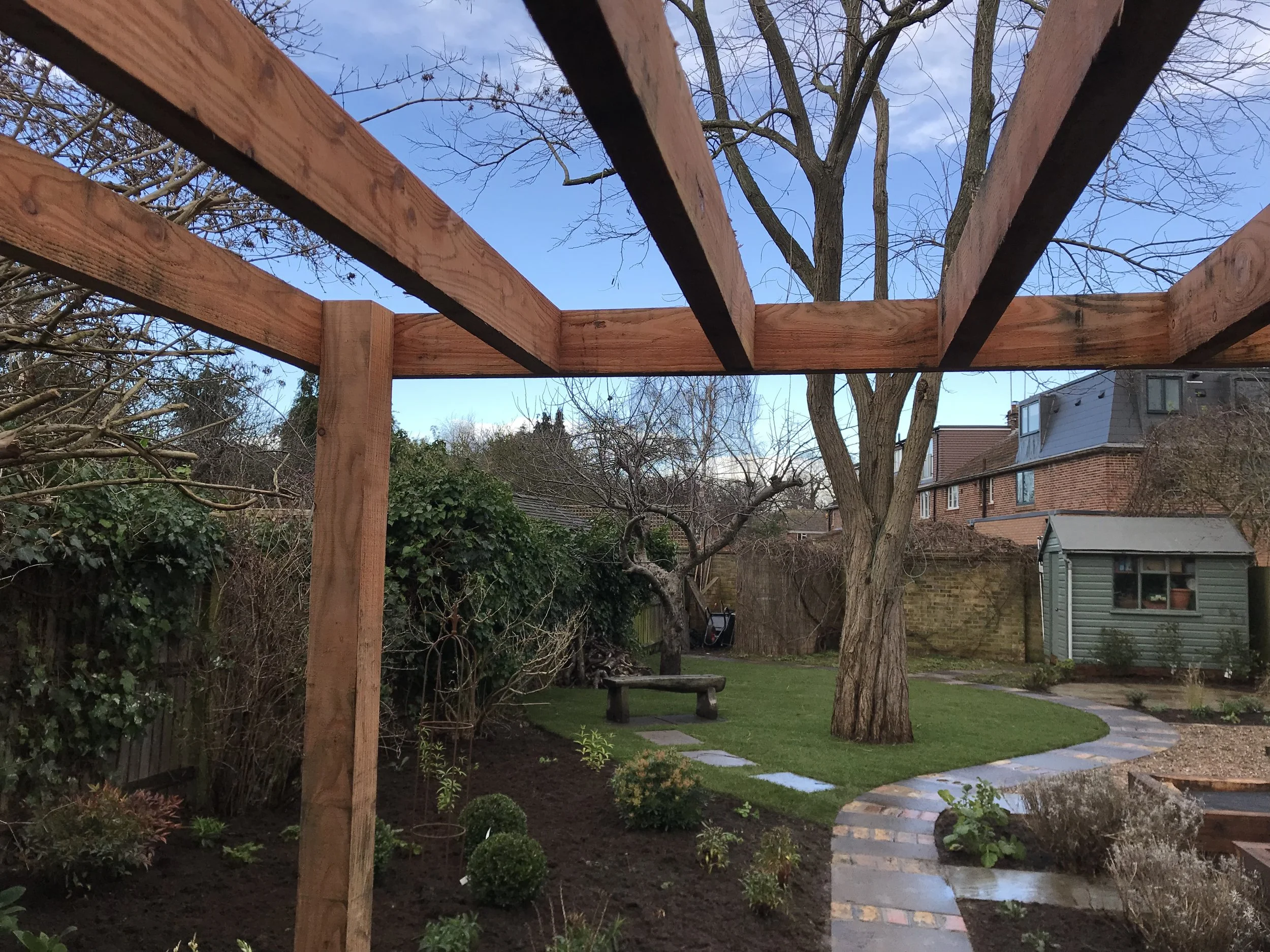 View of a backyard garden during daytime, seen from under construction wooden framework. There are trees, a brick pathway, a small bush, and a green shed, with neighboring houses in the background.