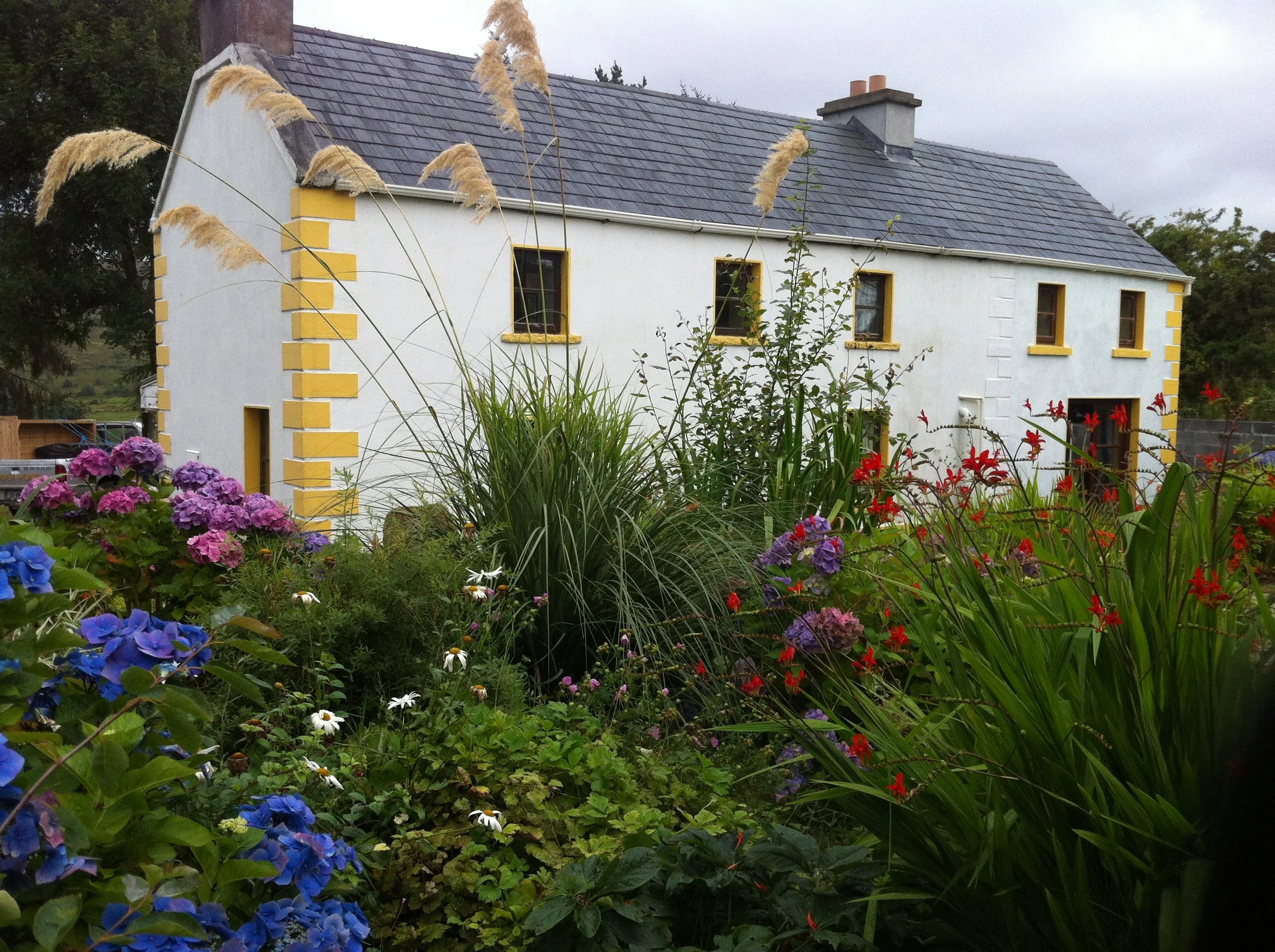 A white house with yellow accents around windows and corners, surrounded by a lush garden with various colorful flowers and tall grasses under an overcast sky.