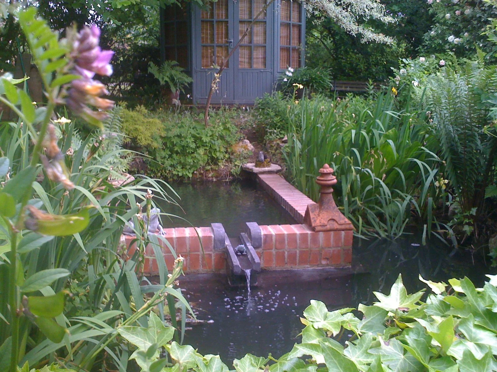A small pond with water flowing from a brick fountain in a lush garden, surrounded by green plants, trees, and a blue shed in the background.