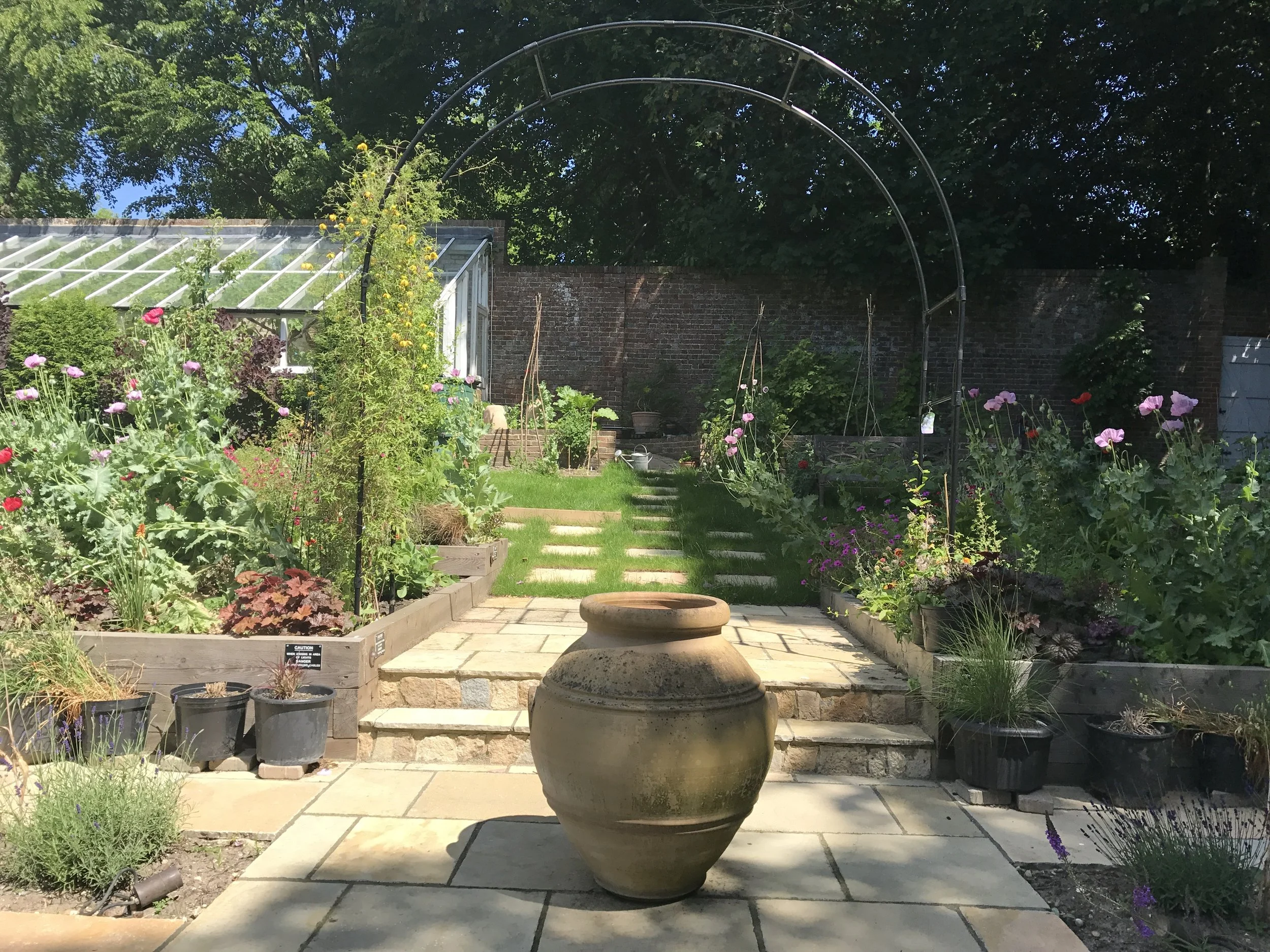 A colorful garden with a stone patio, potted plants, flower beds, a grassy pathway, and a greenhouse in the background.