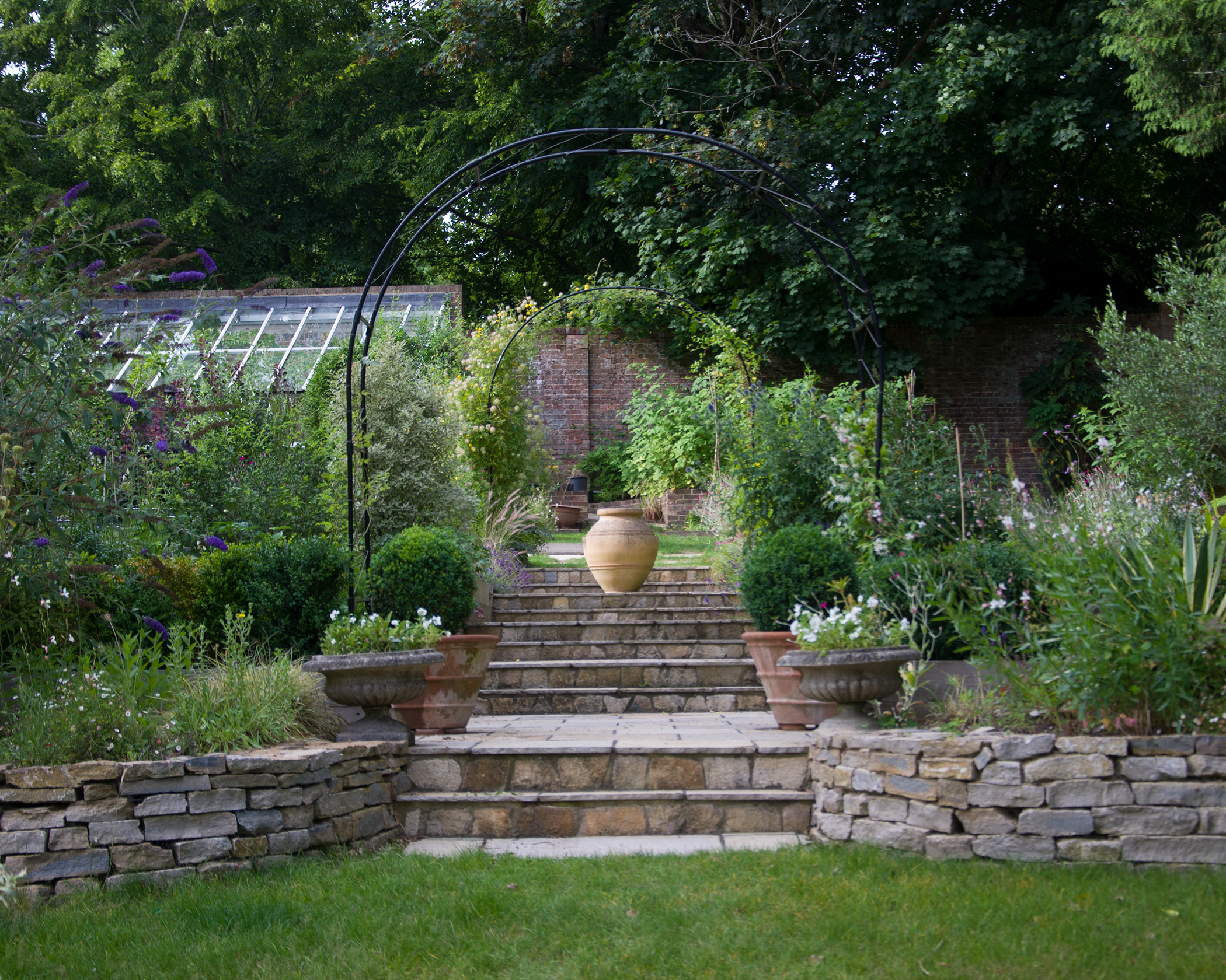 A lush garden with stone steps leading up to a landscaped area with potted plants, arched metal garden trellises, and a variety of flowering plants and greenery.