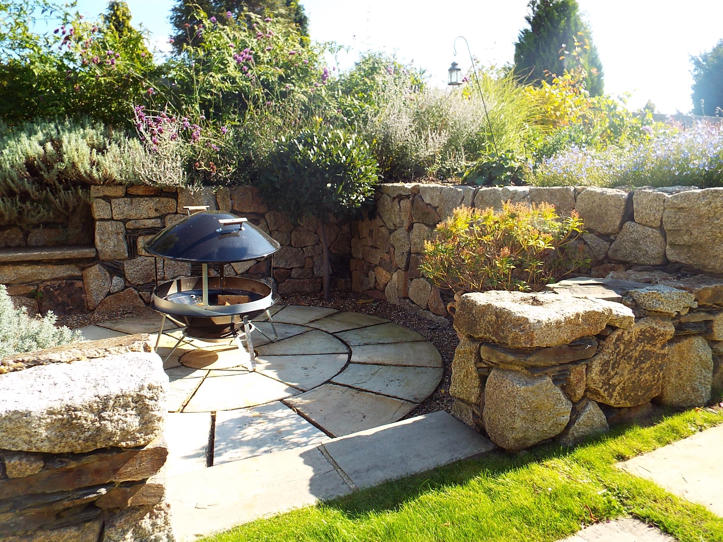 Backyard fire pit area with stone patio, surrounded by a stone wall and lush plants, with sunlight and a lantern overhead.