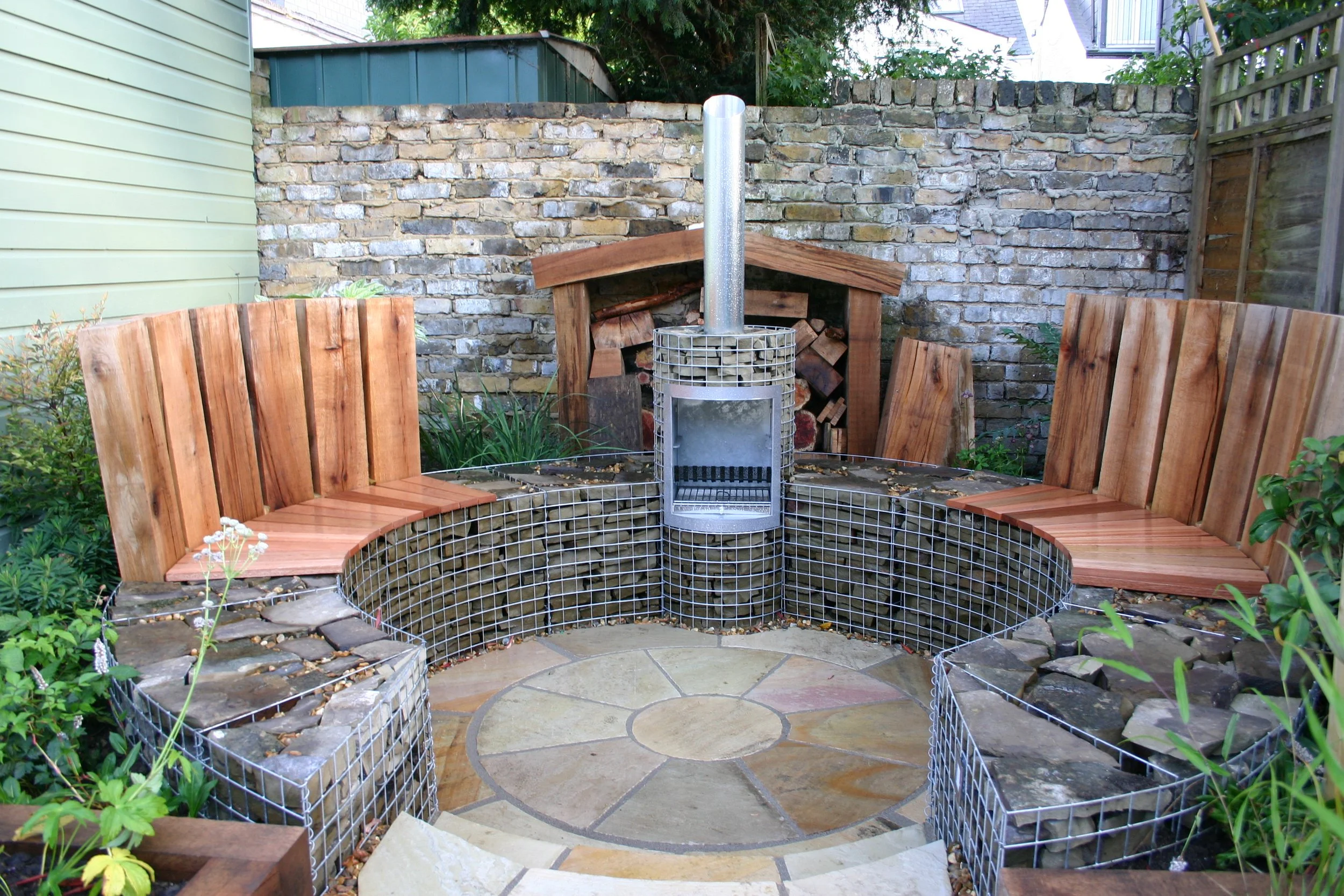 A backyard fire pit with wooden seating, a metal chimney, a brick and stone wall, and a wood storage shed in the background.