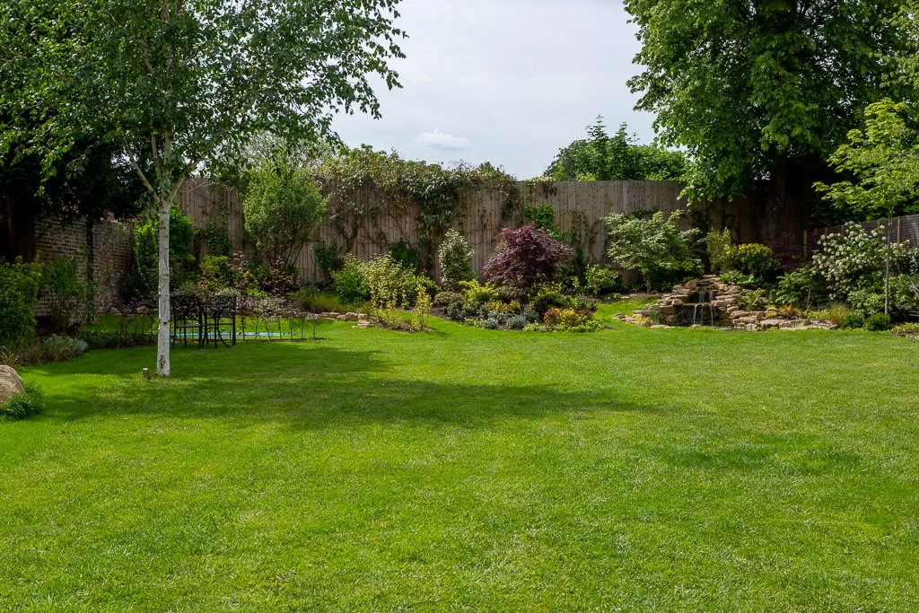 Green backyard with grass, trees, and flower beds, enclosed by a wooden fence, with a small water feature with rocks.