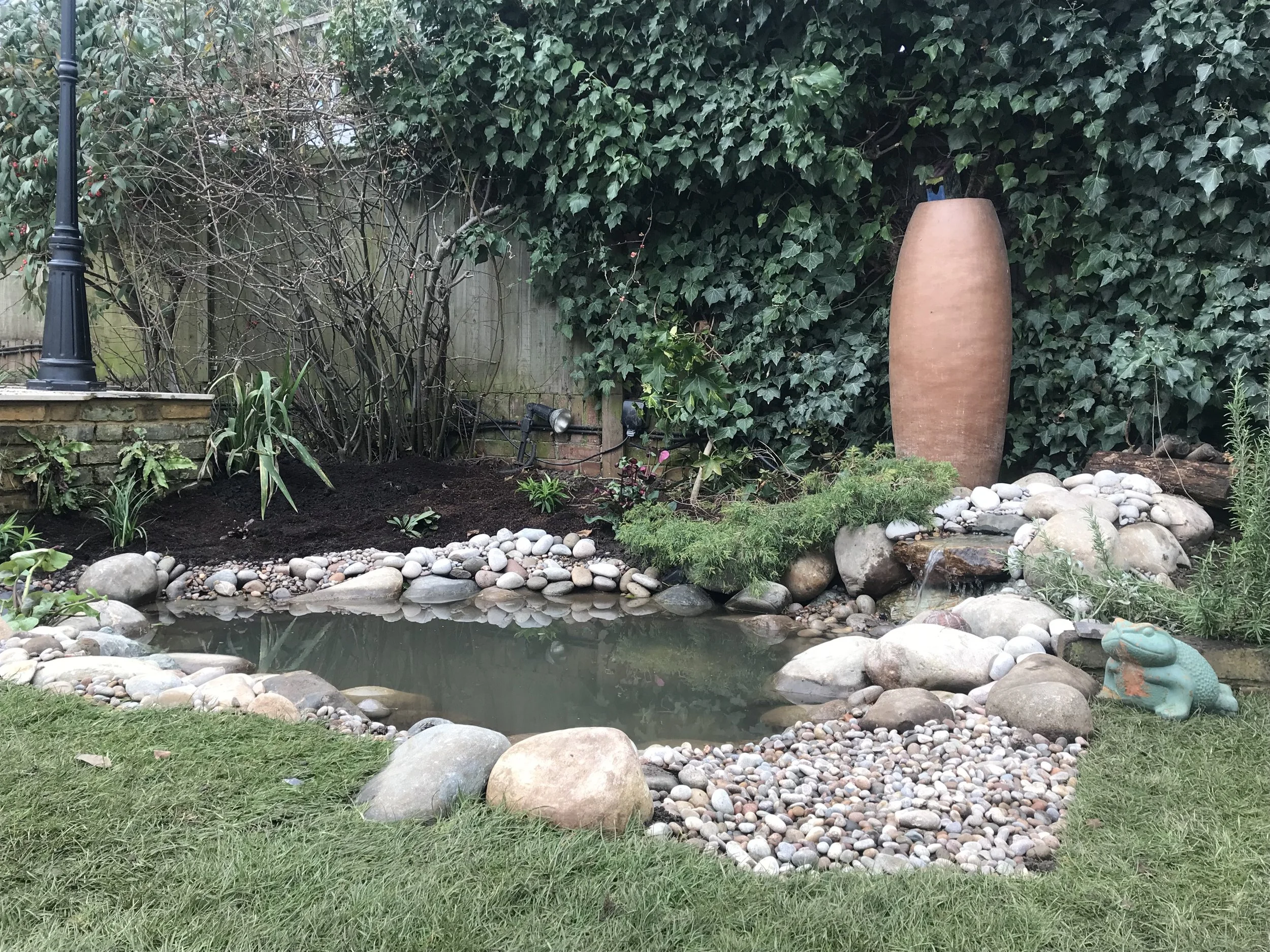 Garden pond surrounded by rocks, with a frog sculpture on the right, tall ceramic vase at the back right, and lush green foliage on the fence and bushes.