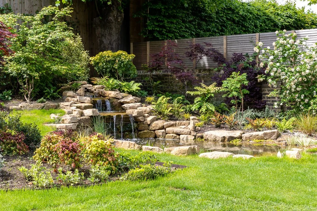A landscaped backyard garden with a rock waterfall, small pond, lush green plants, shrubs, and flowers, enclosed by a wooden fence.