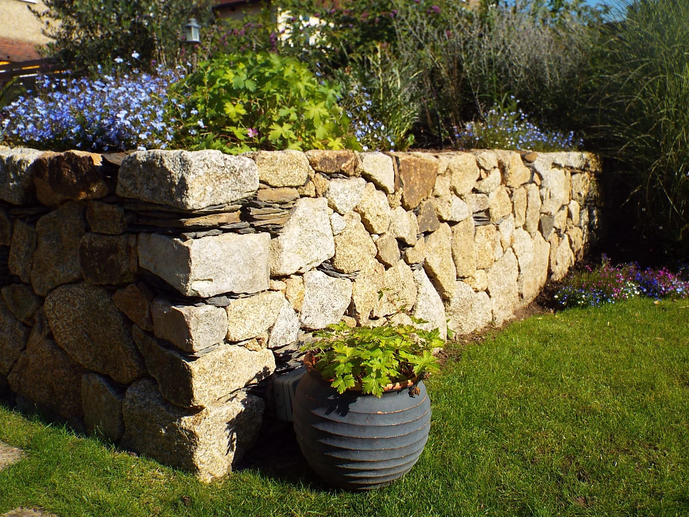 Stone garden wall with potted plant and flowering bushes on a grassy lawn.