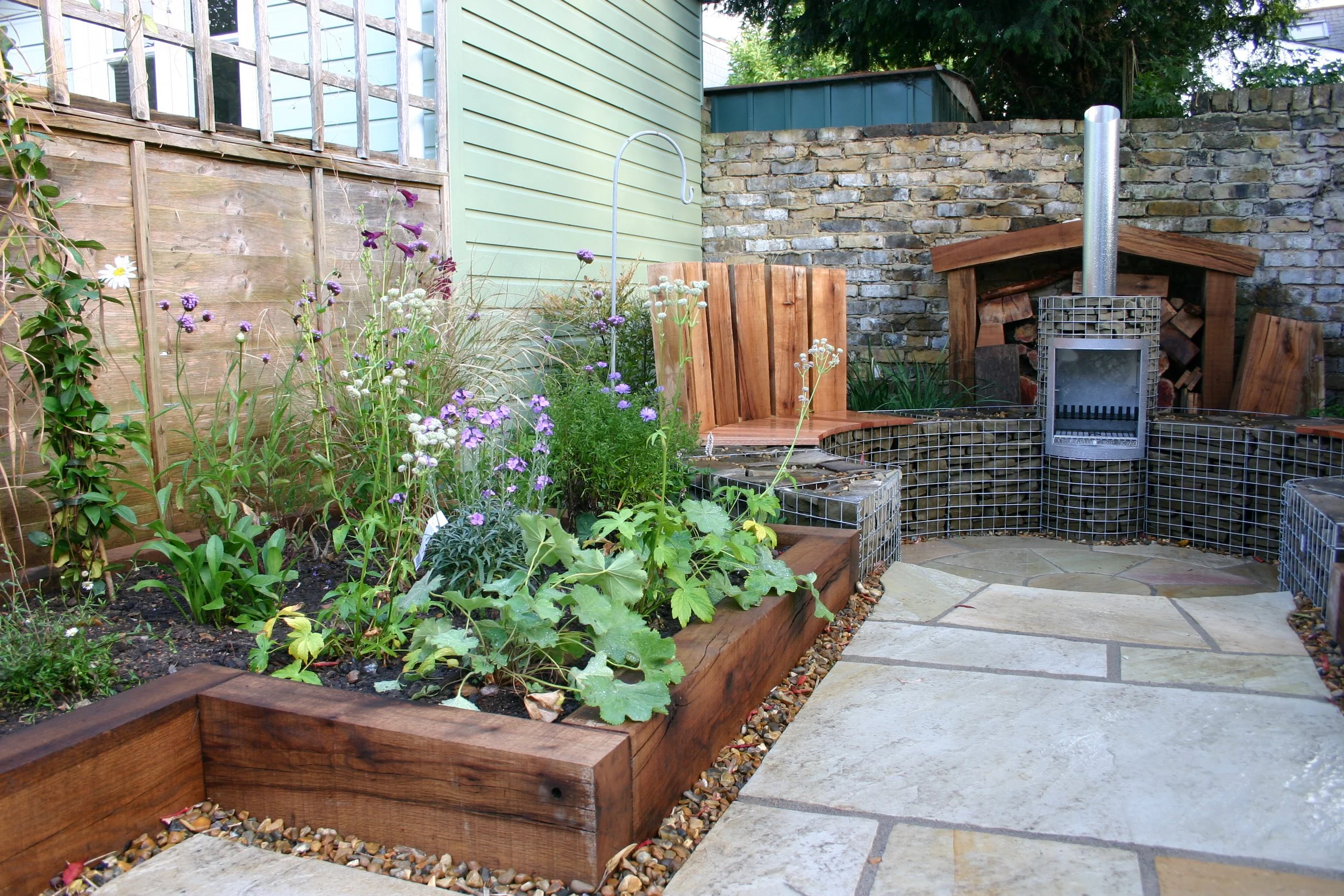 A backyard garden with various plants and flowers, a wooden bench, a metal fireplace with a chimney, and a brick and stone wall.