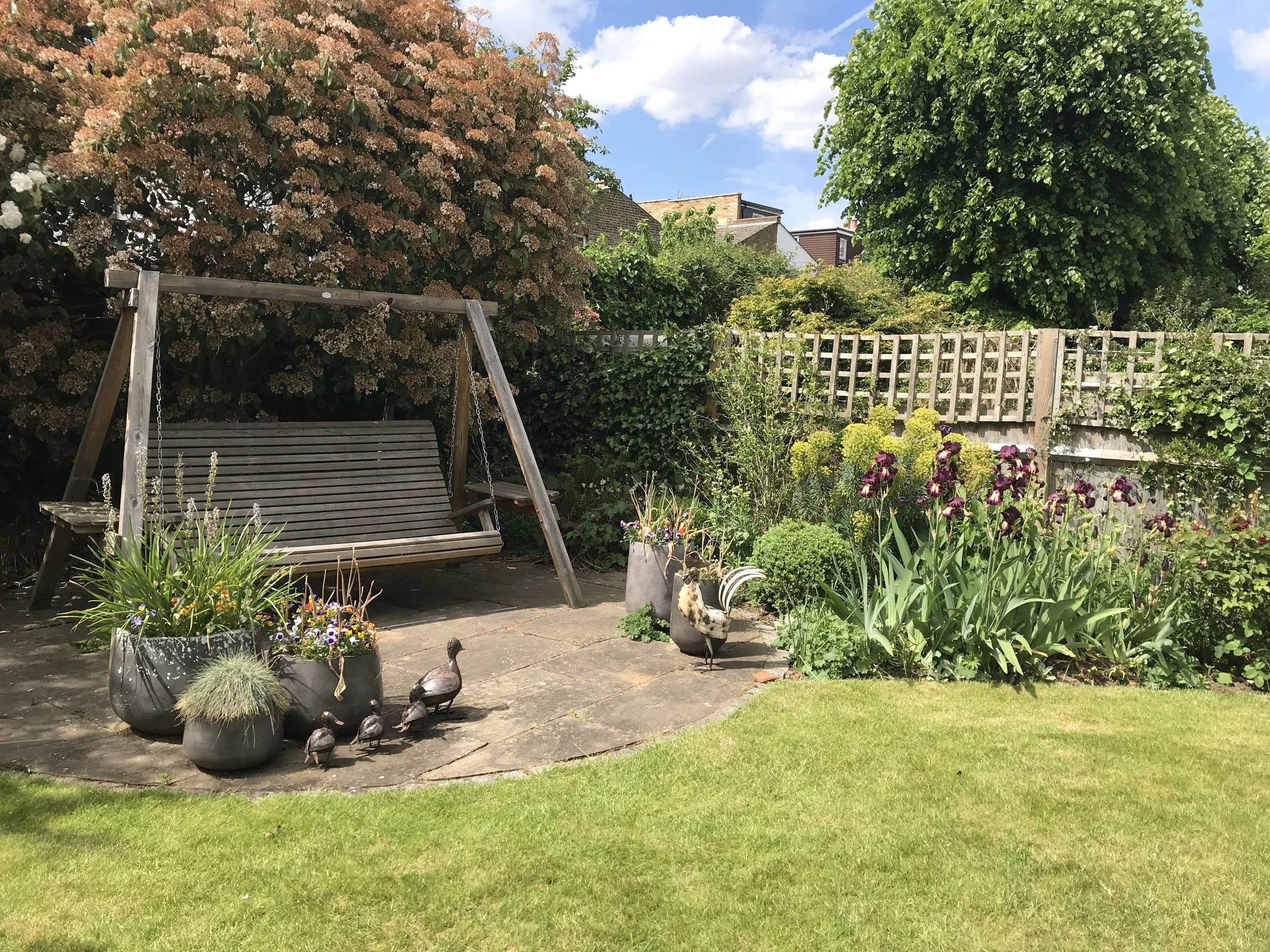 A garden with a wooden swing, potted plants, bird sculptures, blooming flowers, and a green tree in the background, under a partly cloudy sky.