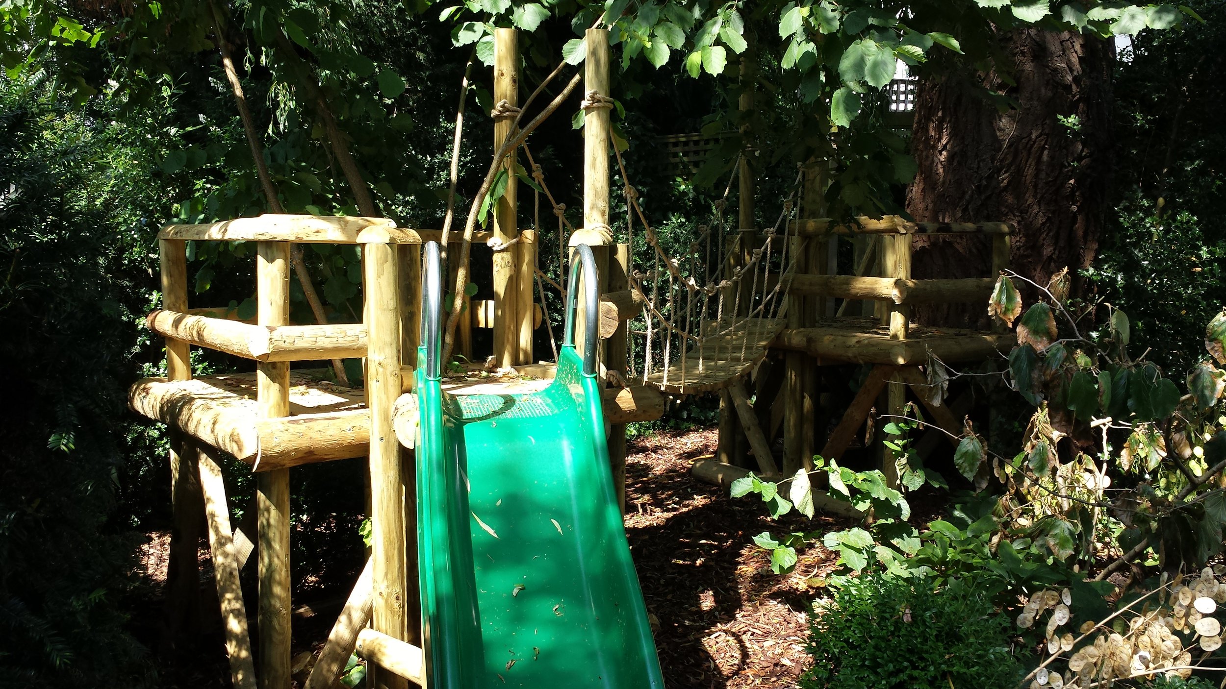 Wooden play structure with a small green slide, rope bridge, and platforms, surrounded by trees and foliage.