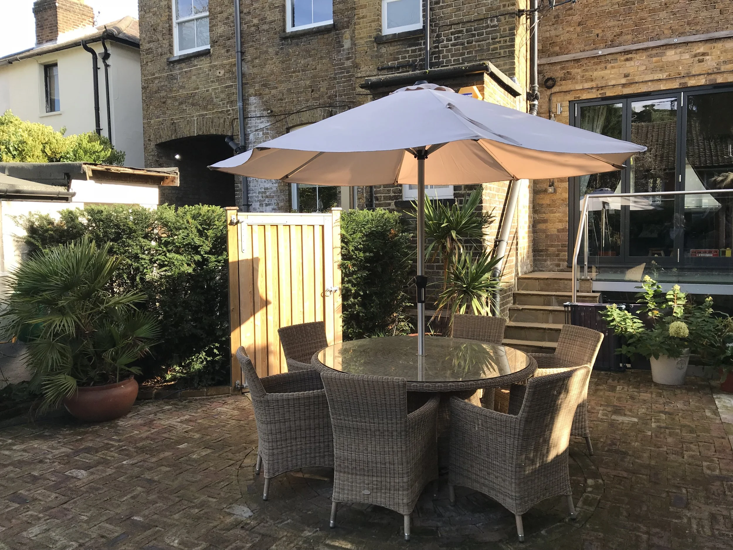 A round glass outdoor table with six wicker chairs around it, under a large beige patio umbrella, on a brick patio. Potted plants and shrubs are nearby, with brick houses and steps in the background.