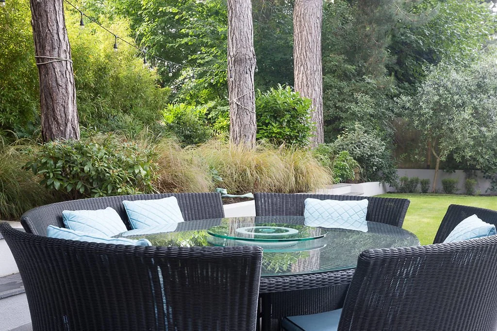 Outdoor patio with a glass table surrounded by black wicker chairs and white pillows, set in a lush garden with trees and bushes.
